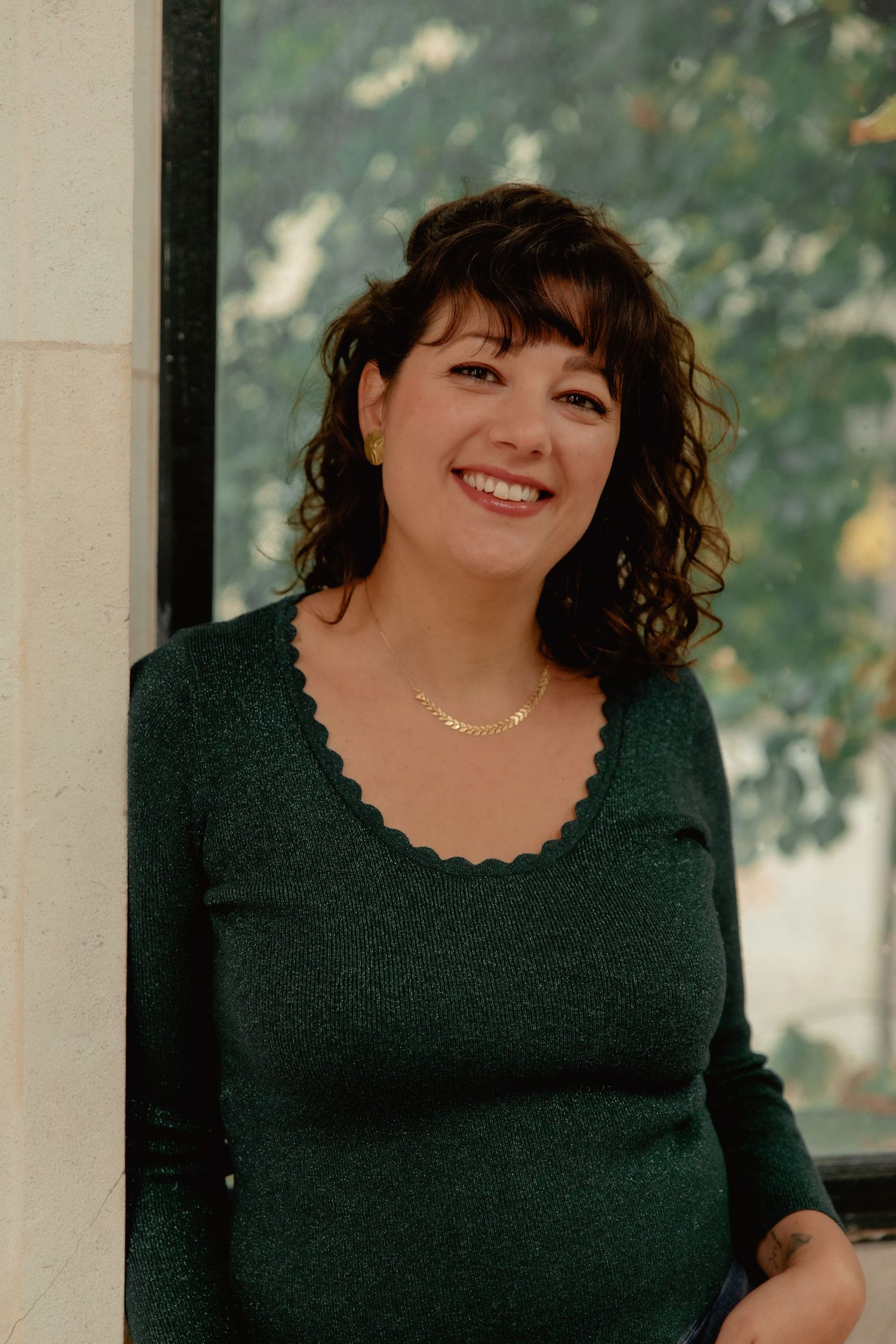A person in a black scoop neck top smiles warmly while posing for a portrait photo outdoors.