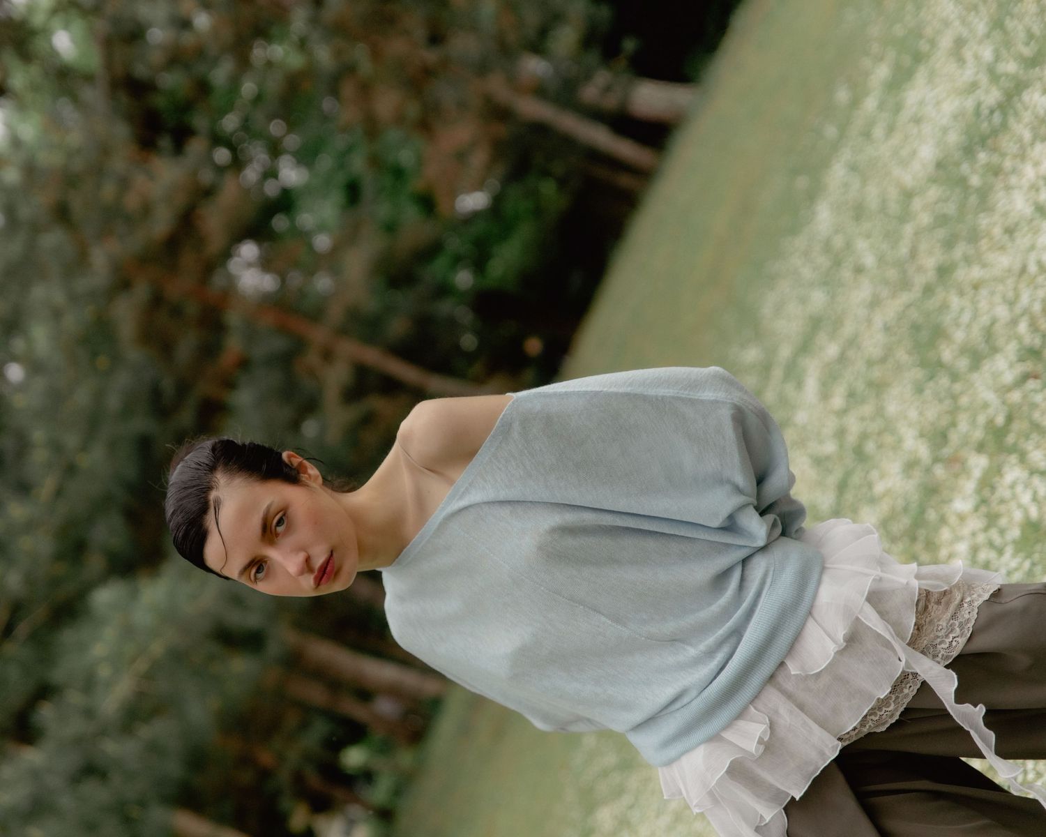 A woman in a light blue sleeveless top and ruffled skirt poses outdoors among lush green foliage.