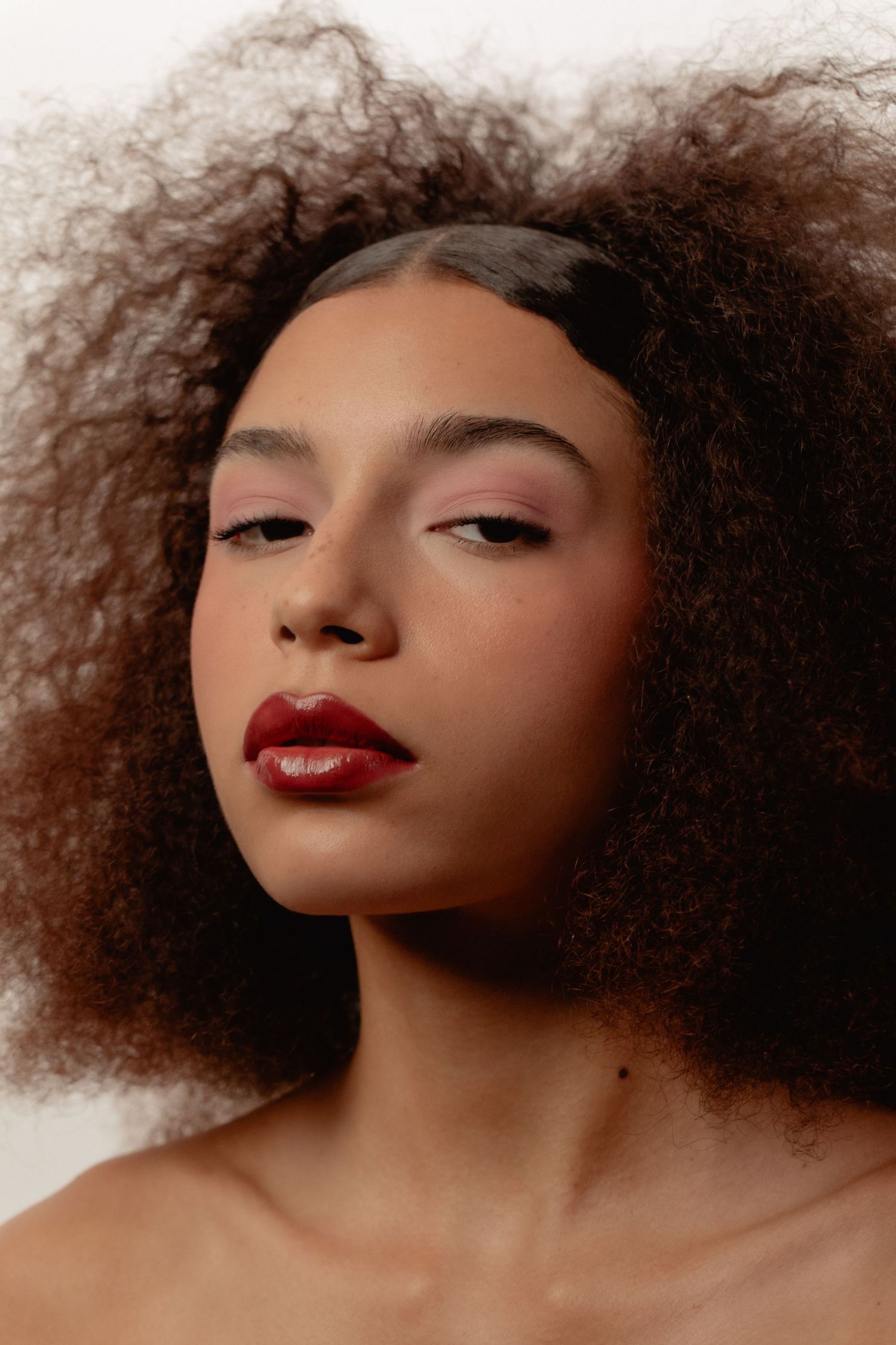 Young woman with voluminous curly hair wearing bold red lipstick and subtle eye makeup against white background.
