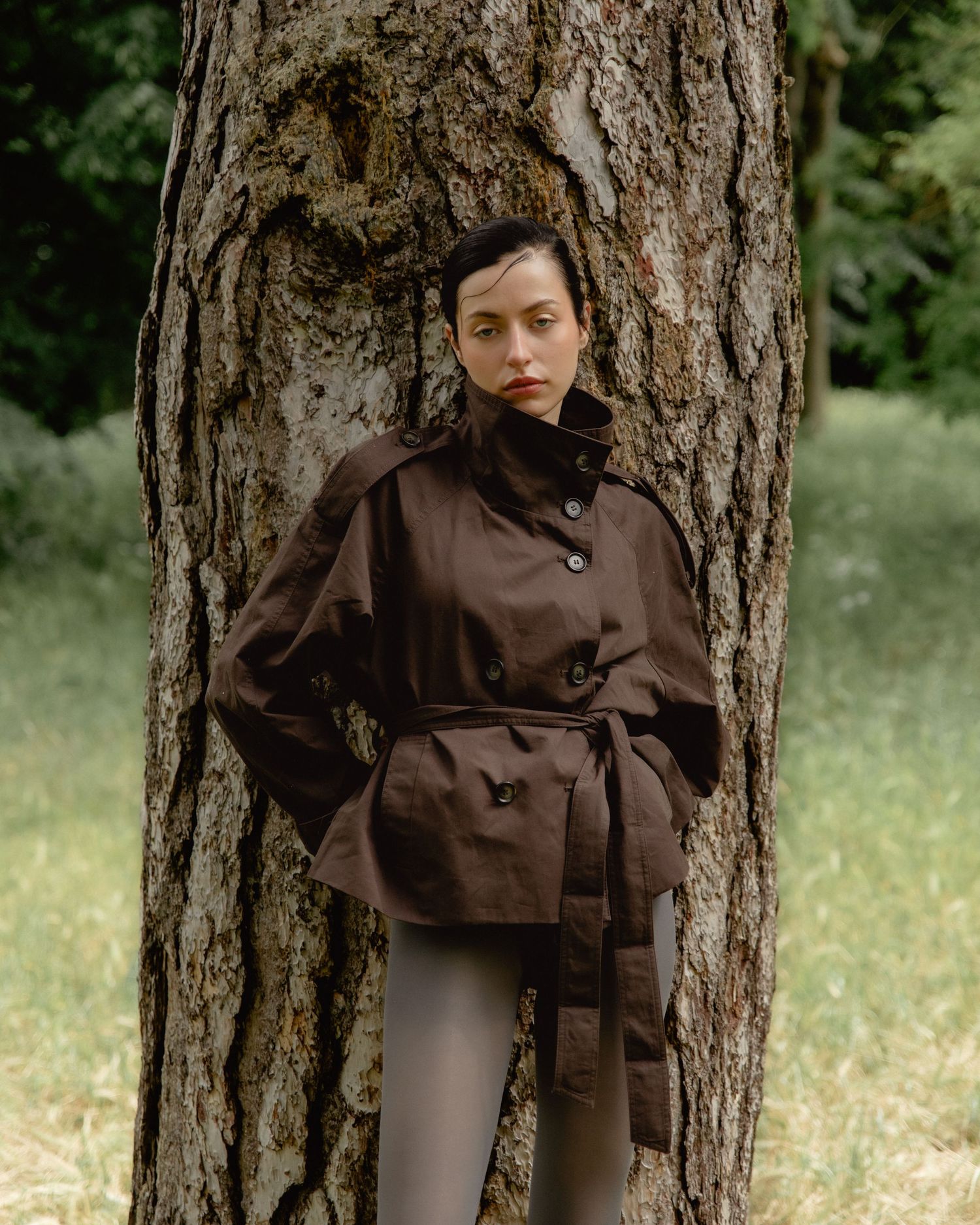 Woman in dark brown belted jacket leaning against a tree trunk in a lush green park setting.