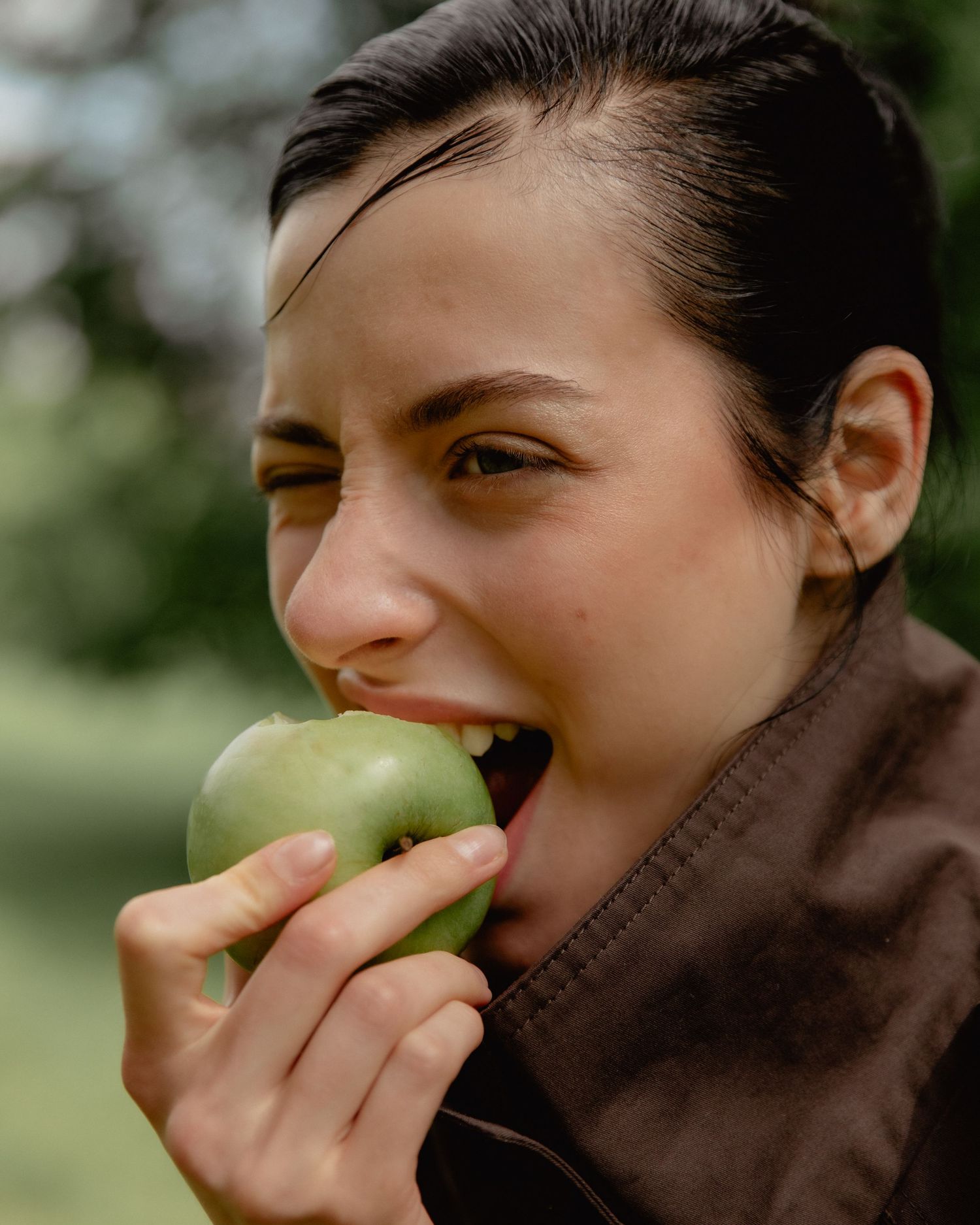 A young woman with dark hair biting into a fresh green apple outdoors, surrounded by lush greenery.