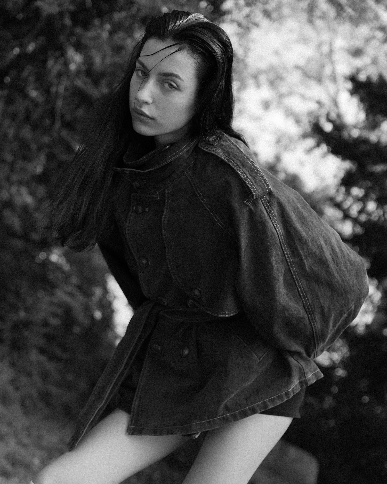 Black and white photo of a young woman in an oversized denim jacket posing outdoors among trees.