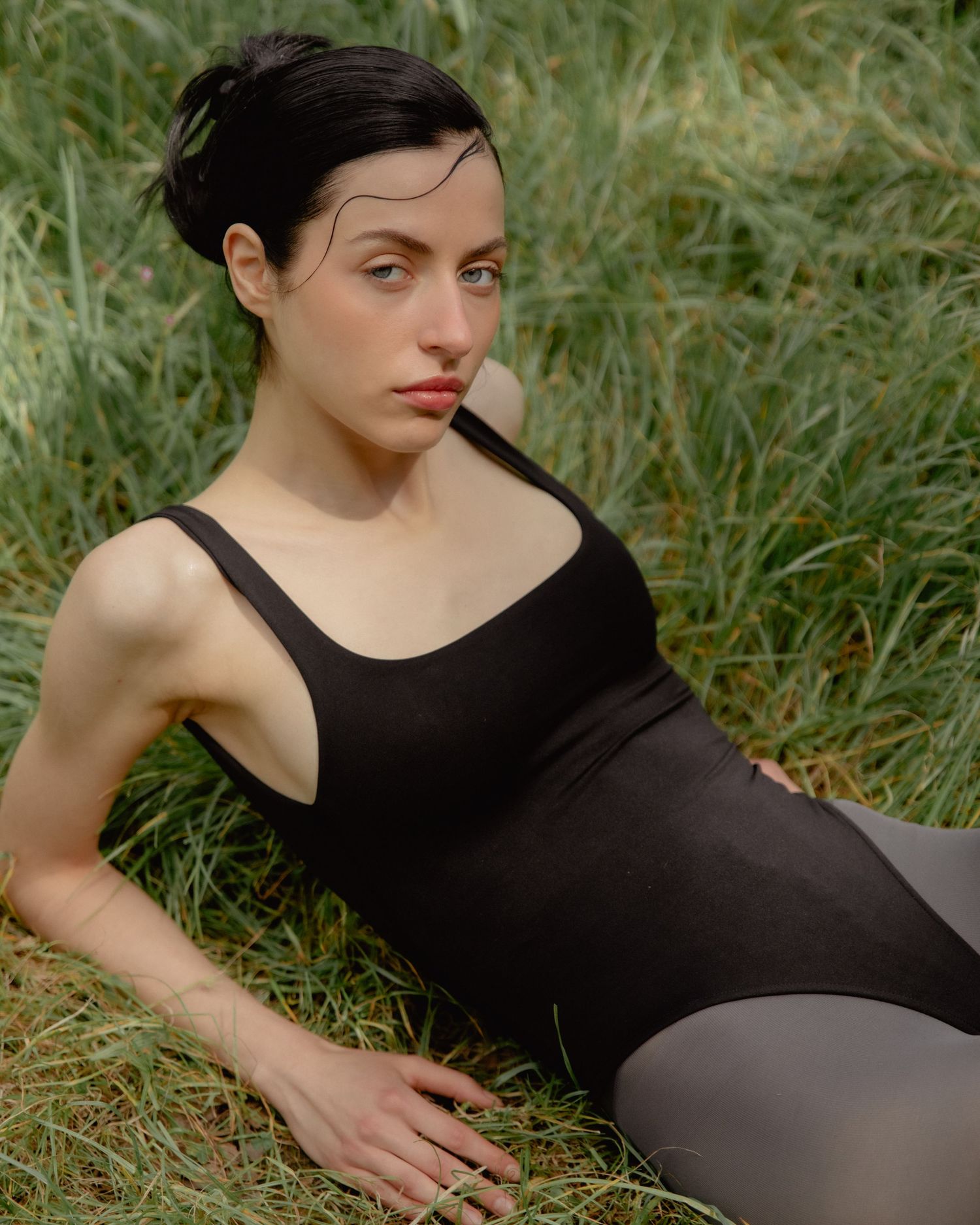 Woman in black bodysuit reclining in green grass, looking over shoulder in natural outdoor setting.