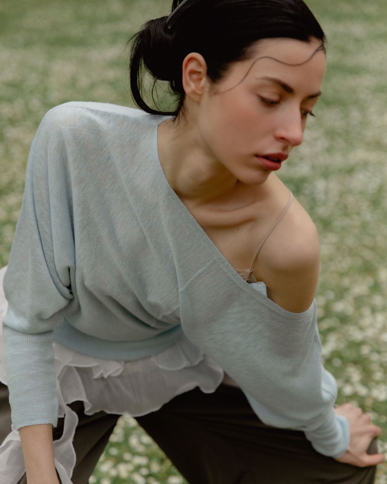 Woman in light blue off-shoulder knit top leaning forward outdoors in a field of white flowers.