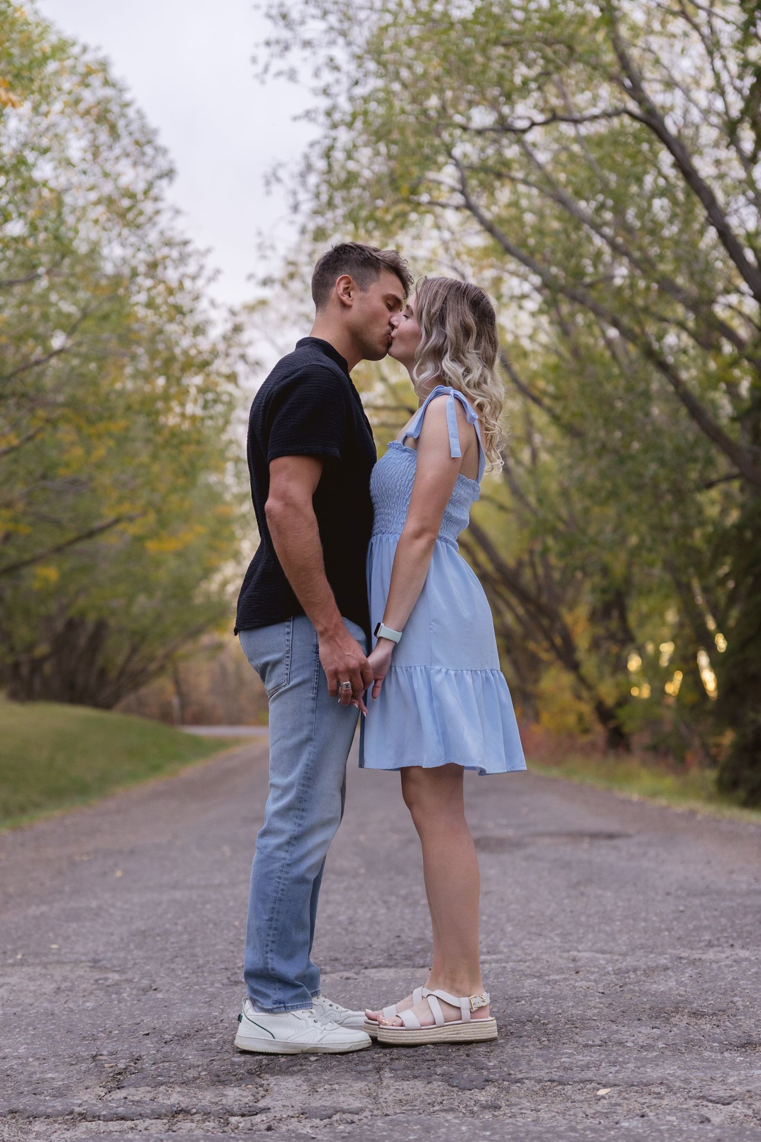 Couple sharing a romantic kiss on a tree-lined path, man in black shirt and jeans, woman in light blue dress.