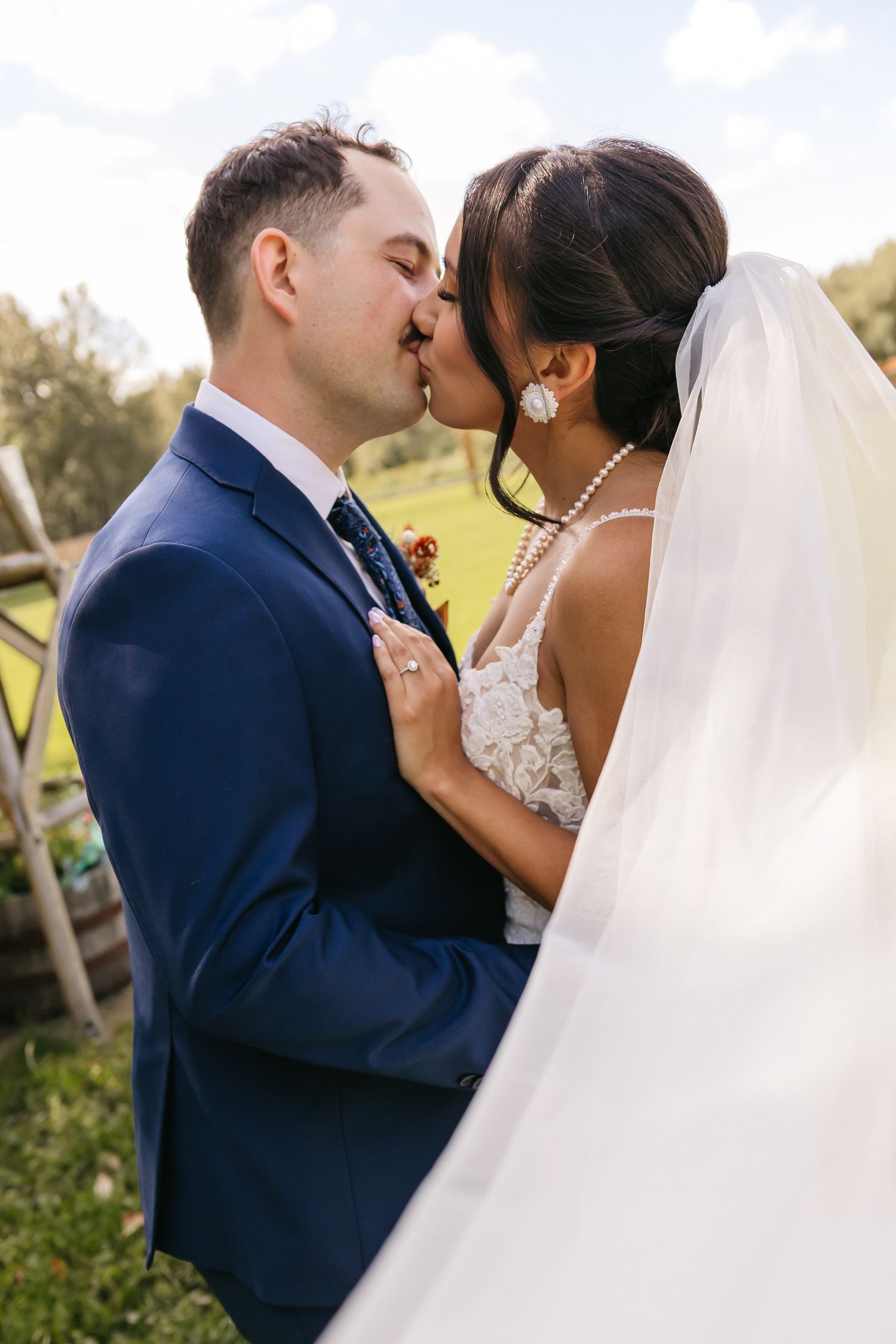 Bride and groom sharing a romantic kiss outdoors, bride wearing white lace gown with veil, groom in navy blue suit.