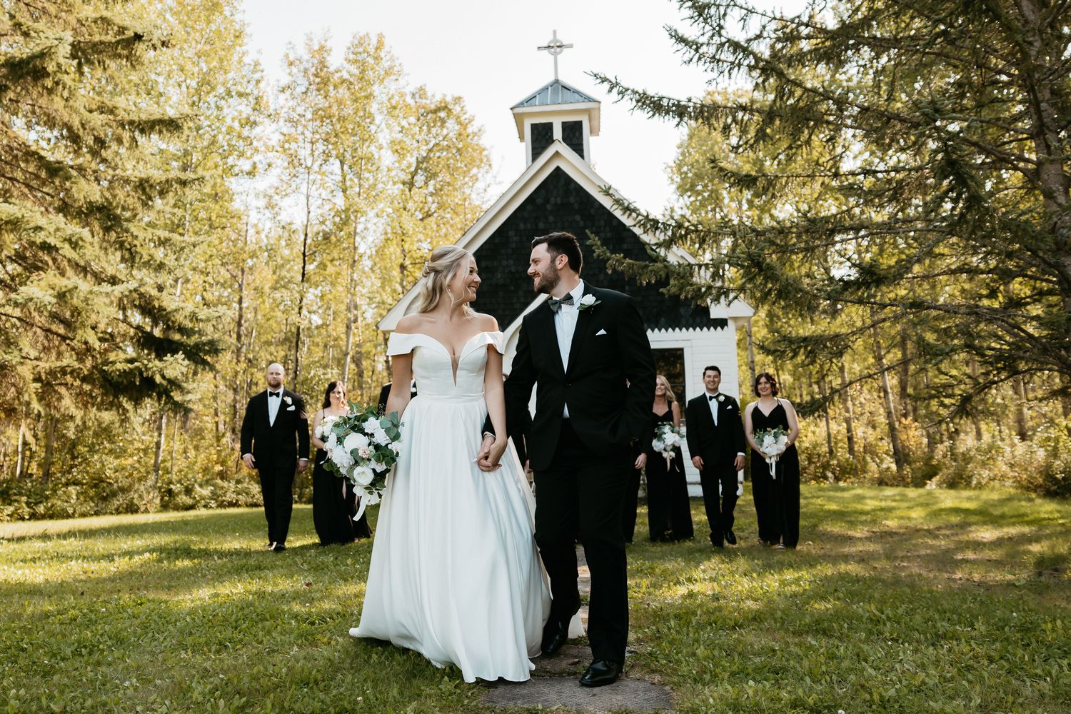 Bride and groom walk hand-in-hand outside a charming chapel surrounded by golden autumn trees with wedding party behind.