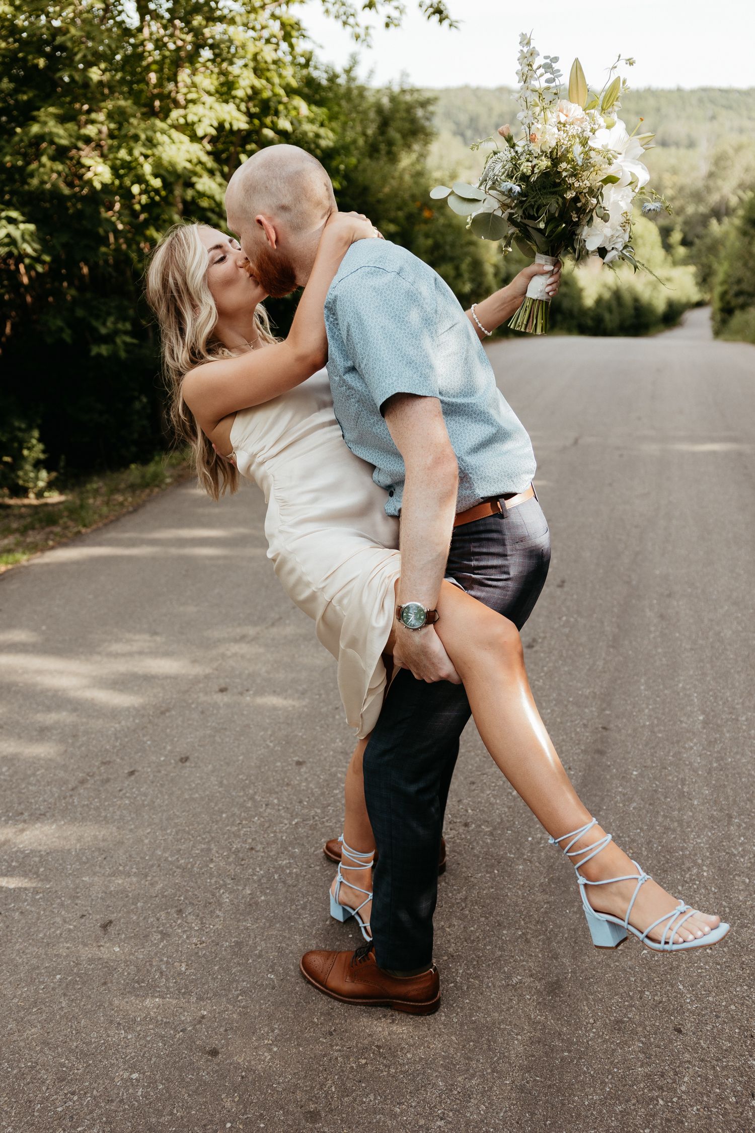 A groom lifts his bride while kissing on a road, she holds a white bouquet, wearing a white dress and blue heels.