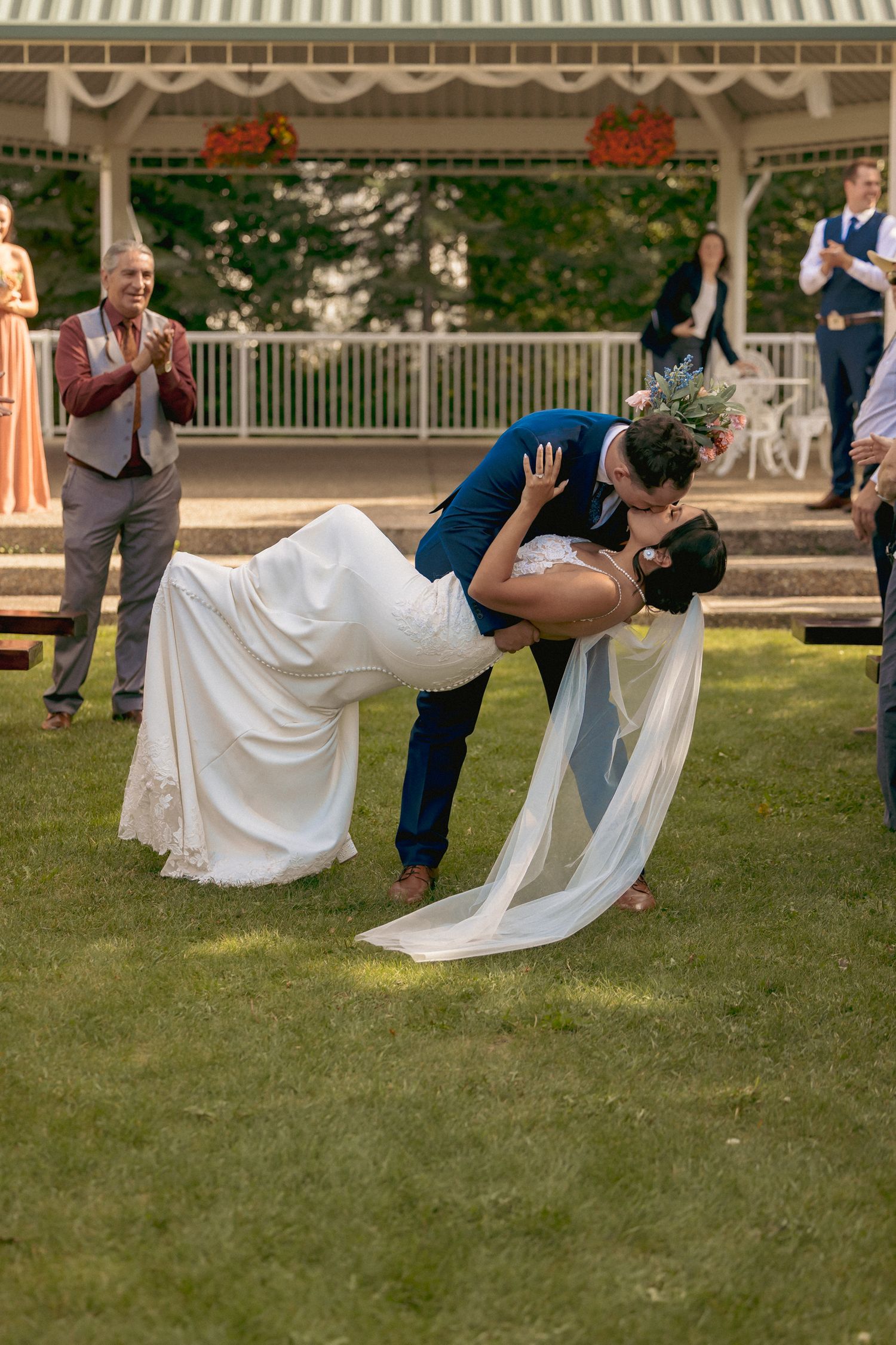 Groom dips bride for a romantic kiss on the lawn during outdoor wedding ceremony with guests applauding in background.