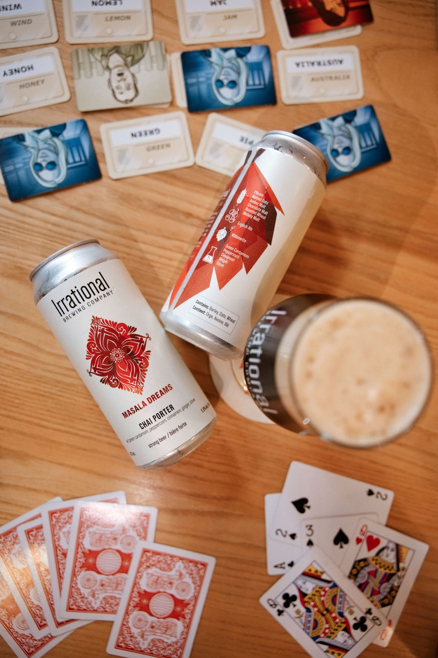 Two Irrational Brewing Masala Dreams Chai Porter cans beside a glass of beer and playing cards on a wooden table.