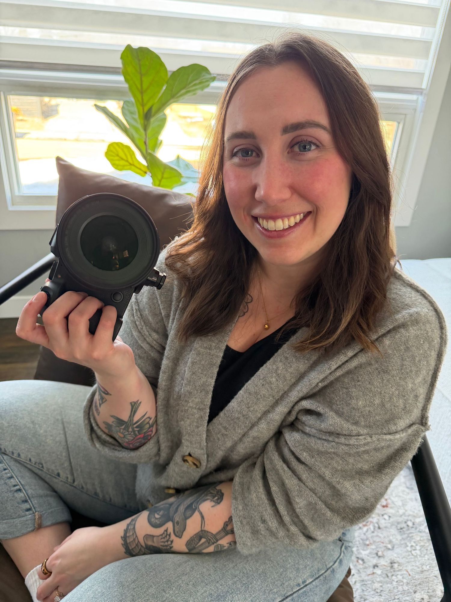 Smiling woman with tattoos holding a camera, wearing a gray cardigan, seated indoors near a window with plants.