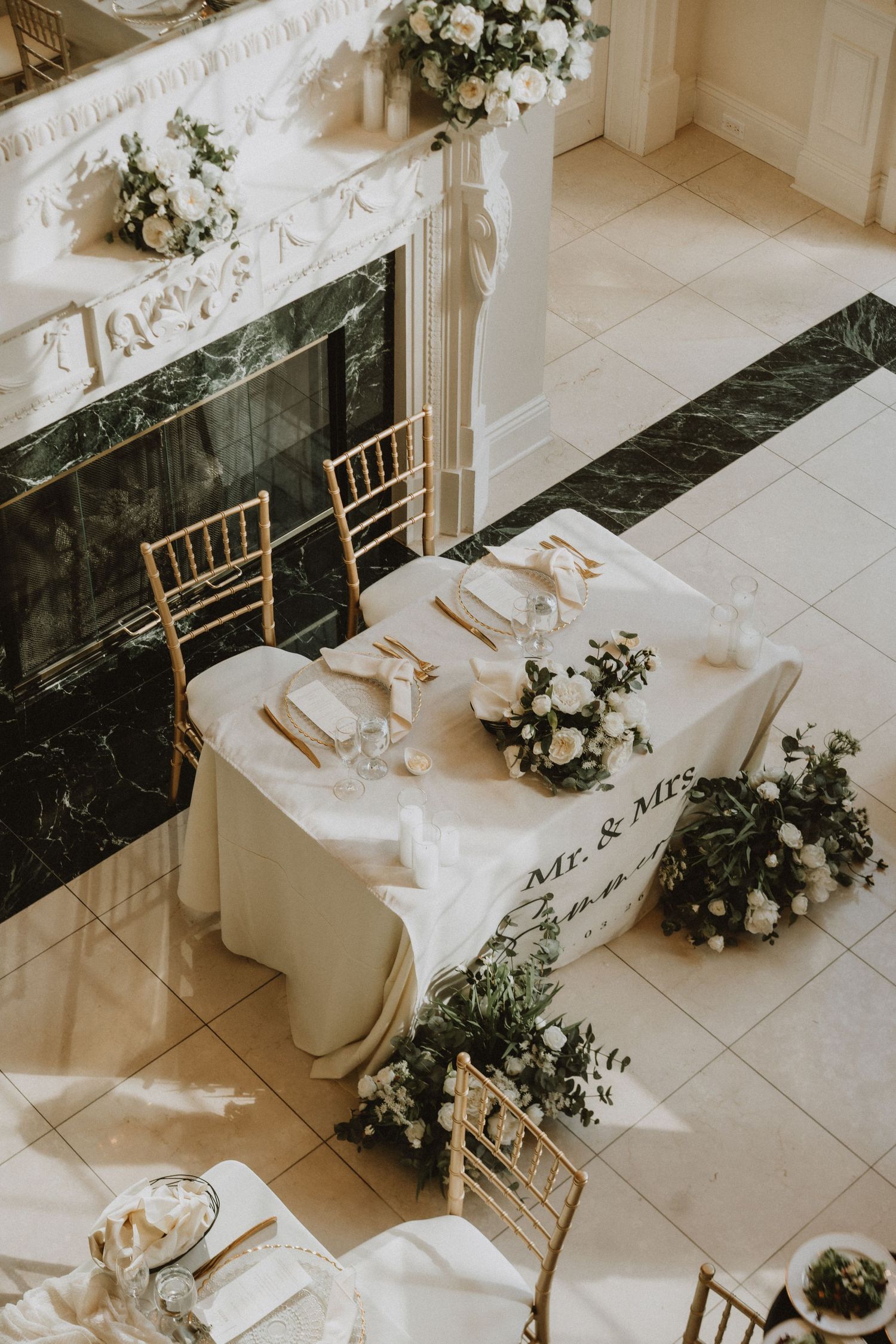 Elegant wedding sweetheart table with white floral arrangements, gold chiavari chairs, and Mr. & Mrs. signage near a marble fireplace.