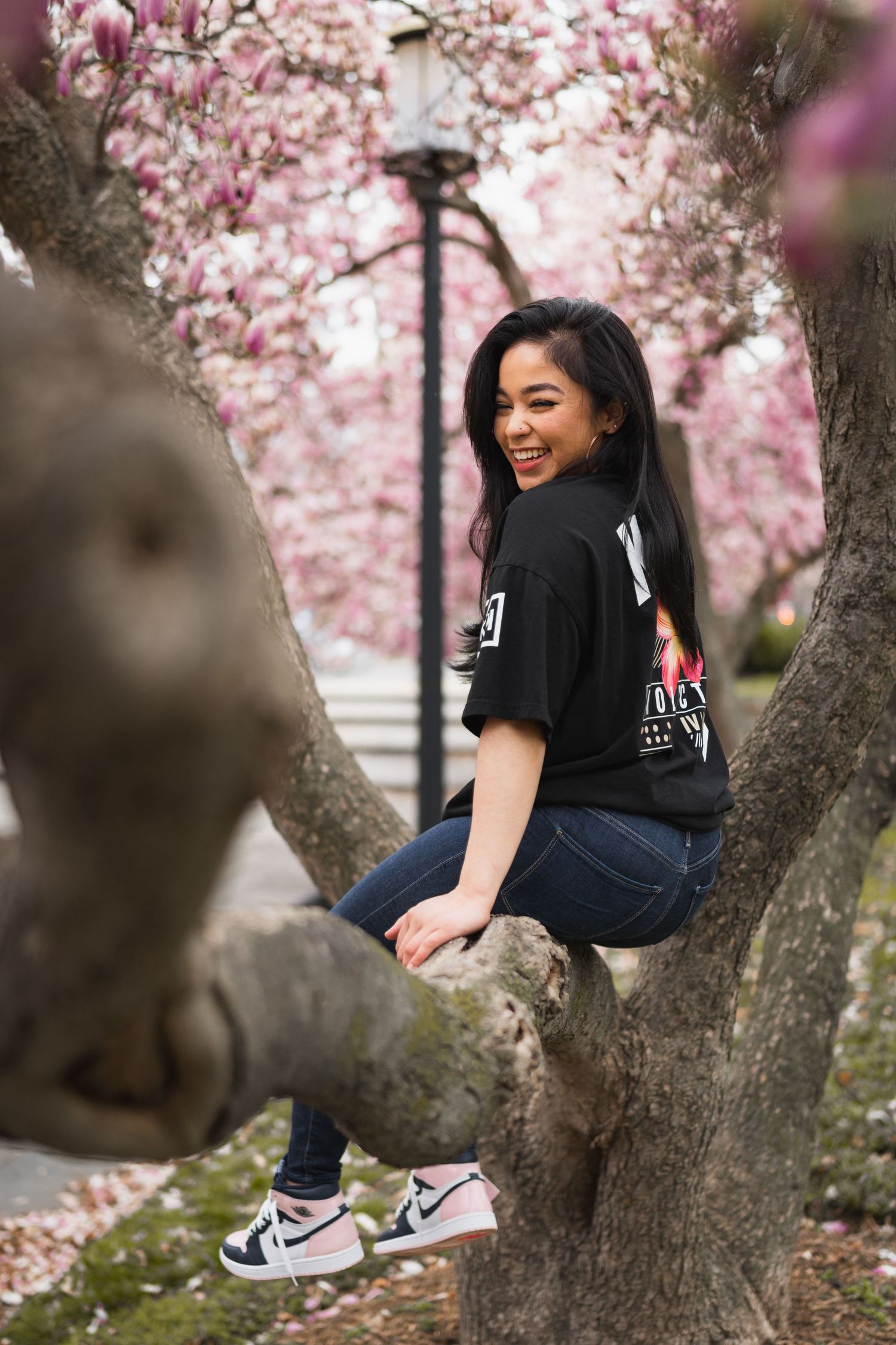 Young woman smiling while sitting in a blooming pink magnolia tree wearing a black graphic tee and Nike Air Jordans.