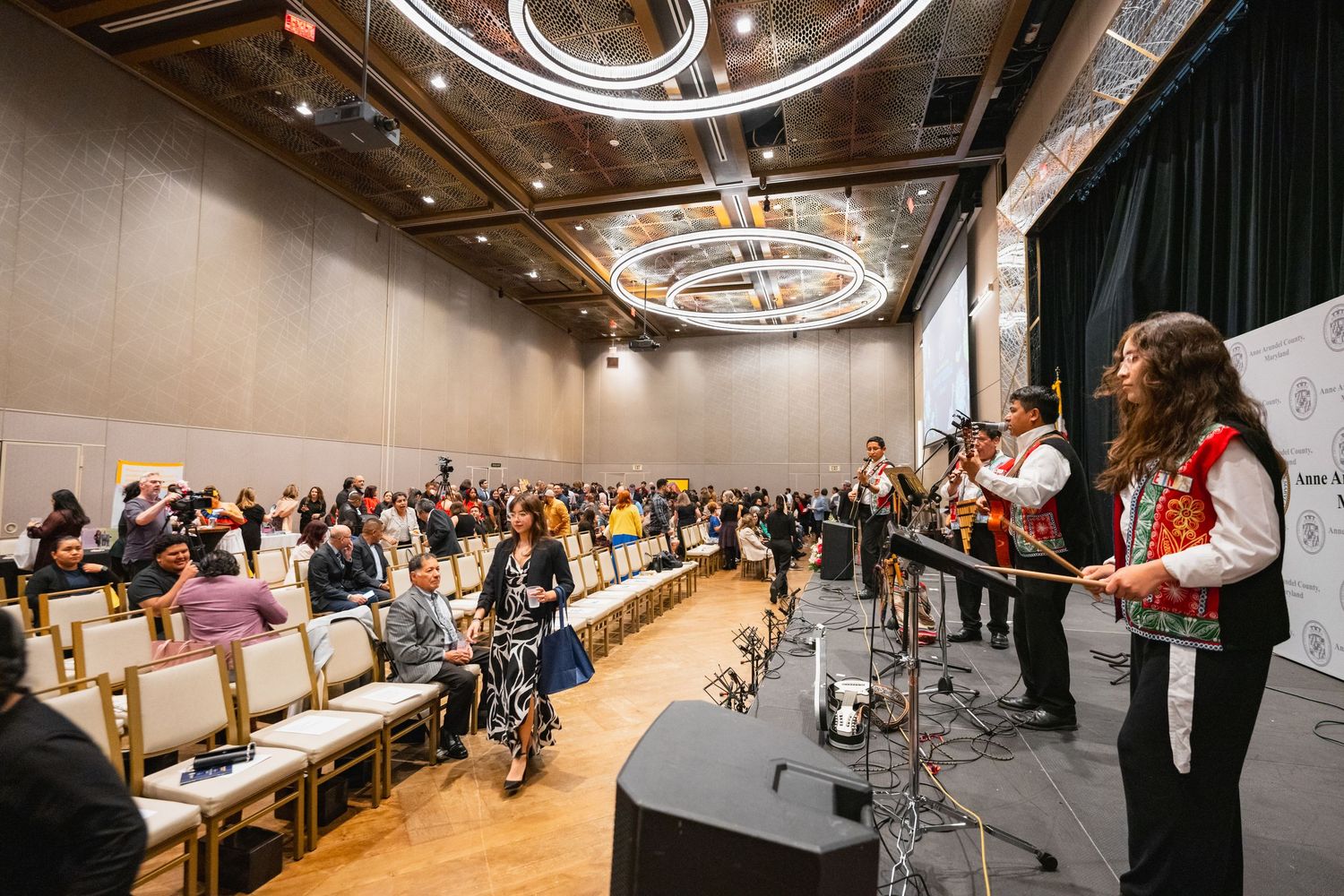 A lively band performs for a large audience in an elegant ballroom with a stunning chandelier overhead.