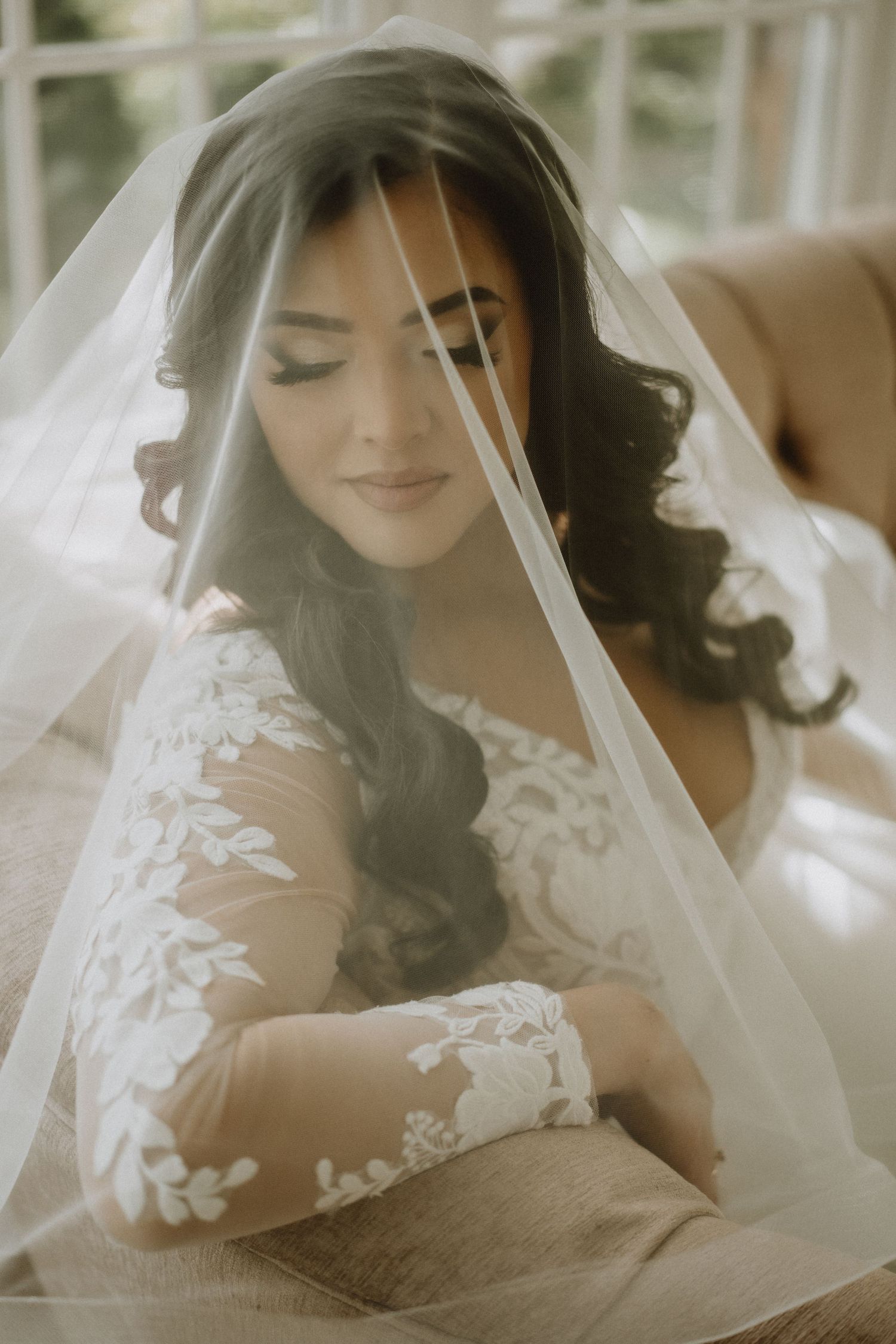 Beautiful bride with dark curly hair draped in a sheer veil wearing a lace long-sleeve wedding gown.