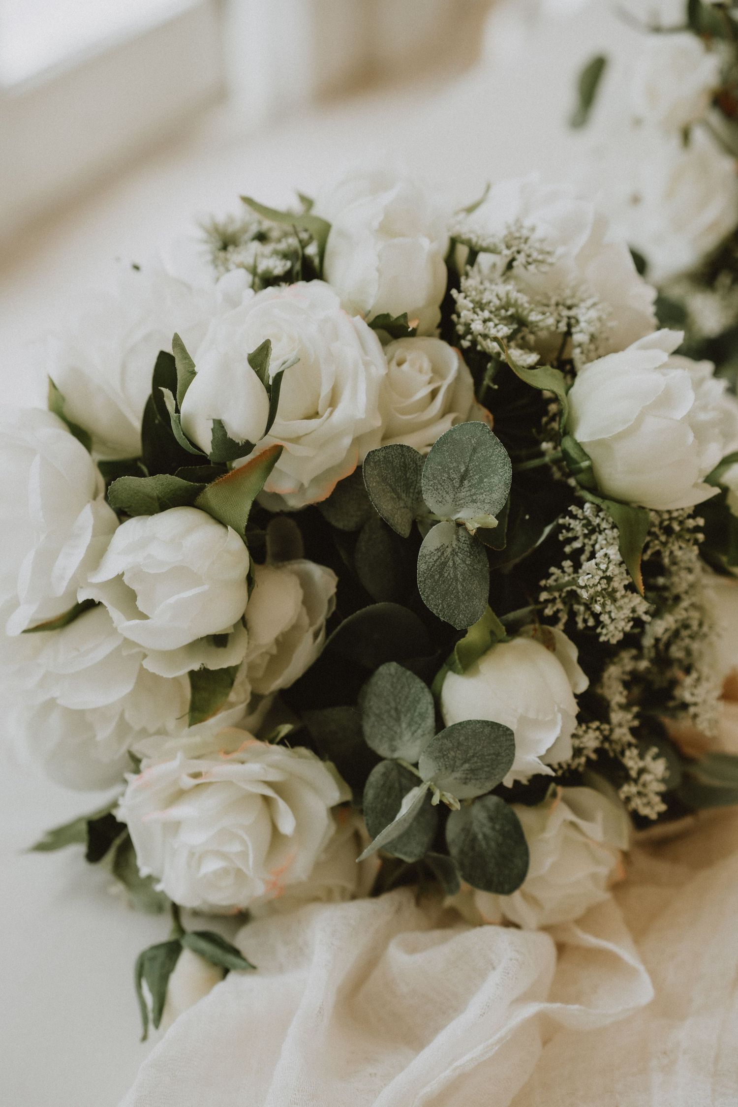 Bridal bouquet of white roses and eucalyptus leaves resting on soft white fabric in natural window light.