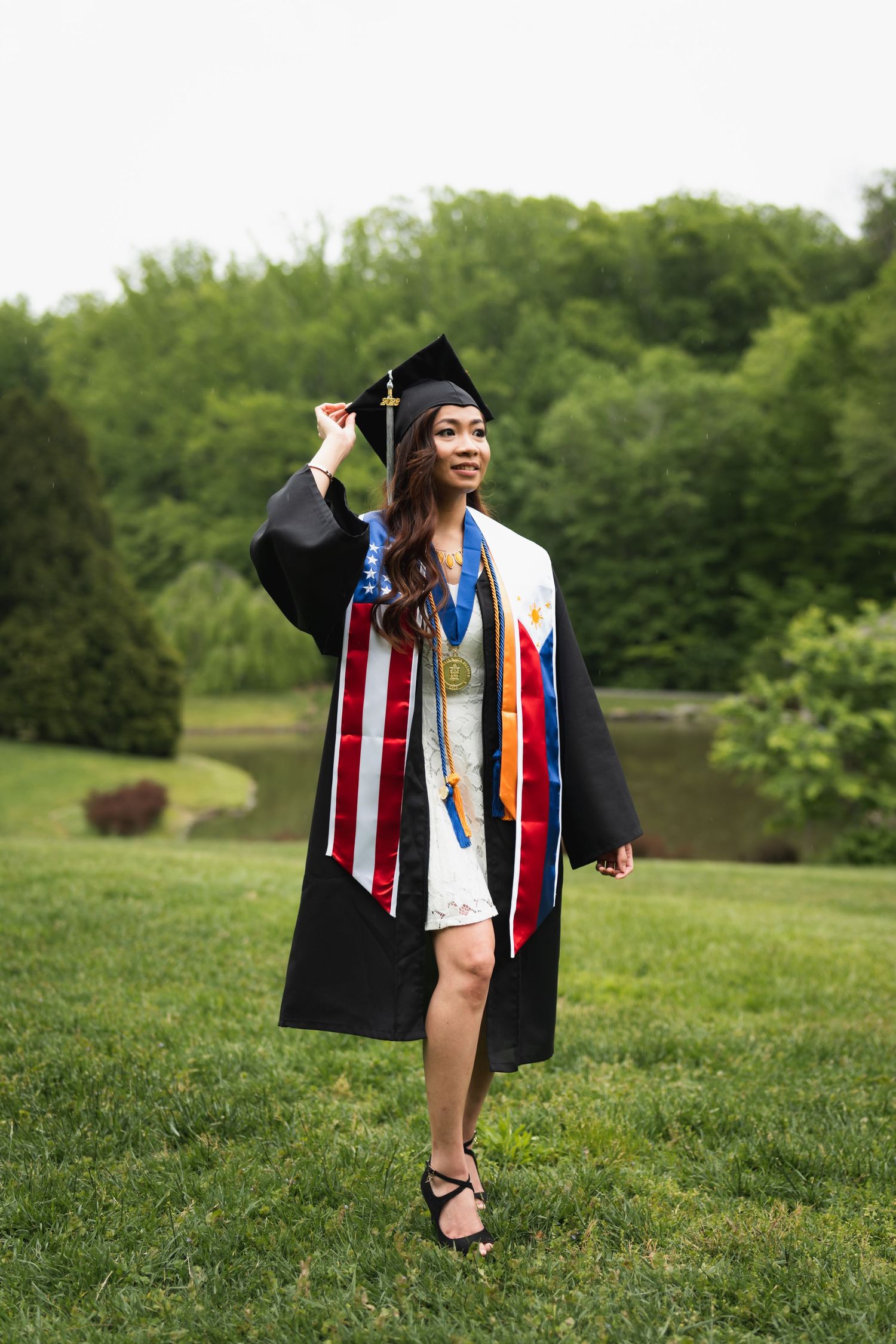 Graduate in black cap and gown wearing an American flag sash and Filipino flag stole in a green park.
