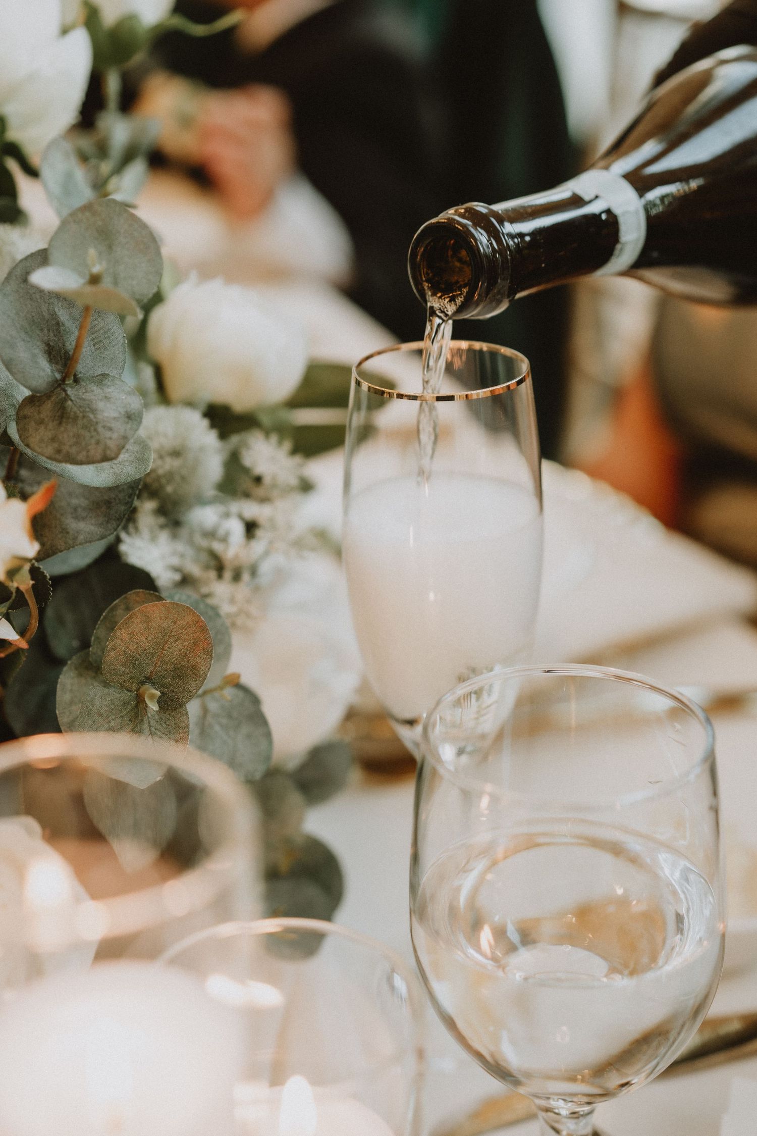Champagne being poured into a flute glass at a wedding reception table with white floral centerpiece and candles.