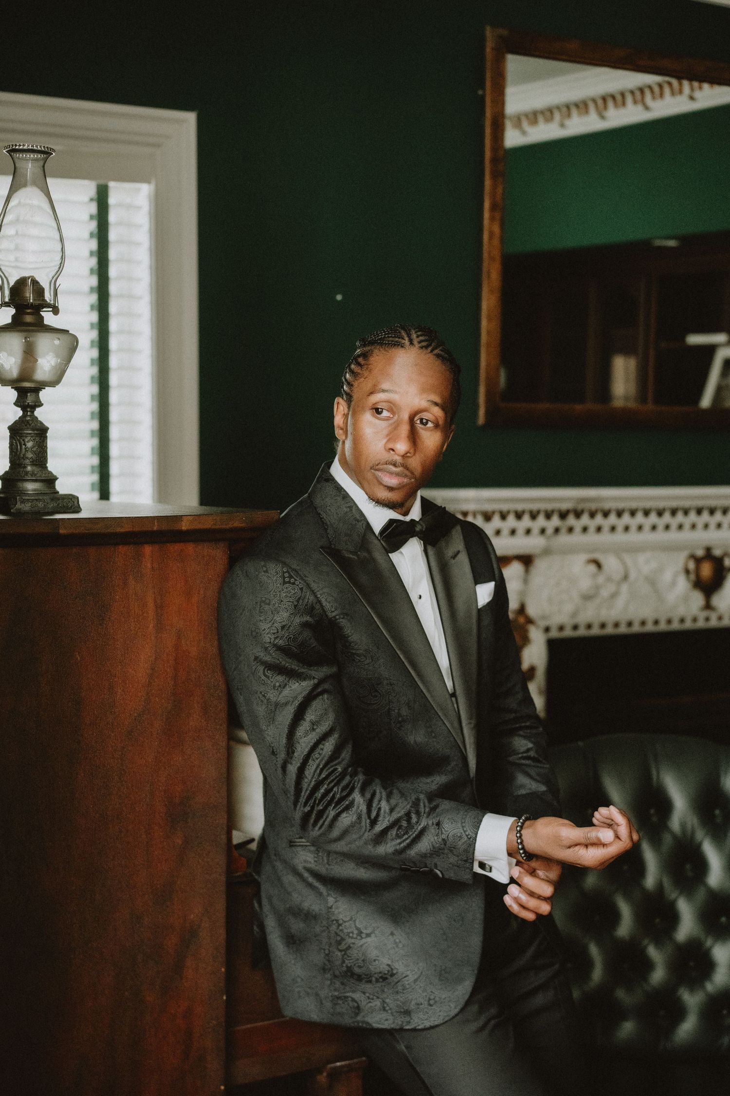Groom in a black floral tuxedo with bow tie seated beside a fireplace in an elegant dark green room.