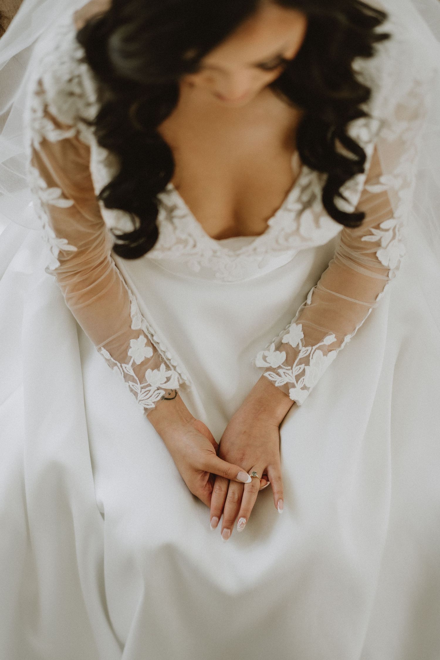 Close-up of bride in lace long-sleeve wedding gown with hands folded showing diamond engagement ring.