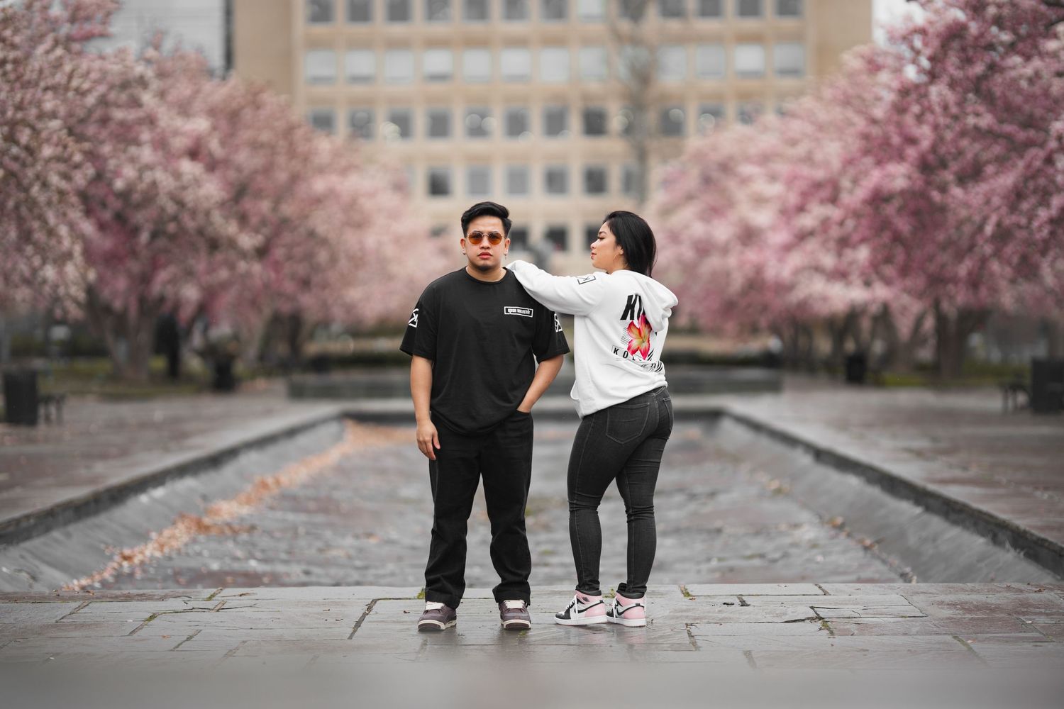 Couple posing between rows of blooming cherry blossom trees near a reflective pool in an urban park.