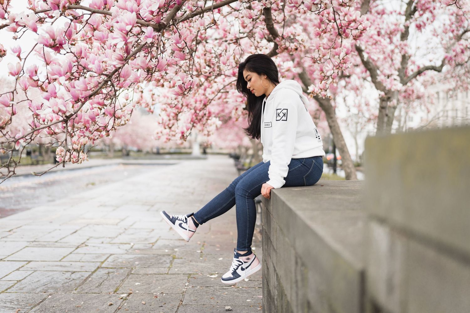 Young woman in a white hoodie sitting on a stone wall beneath blooming pink magnolia trees in spring.
