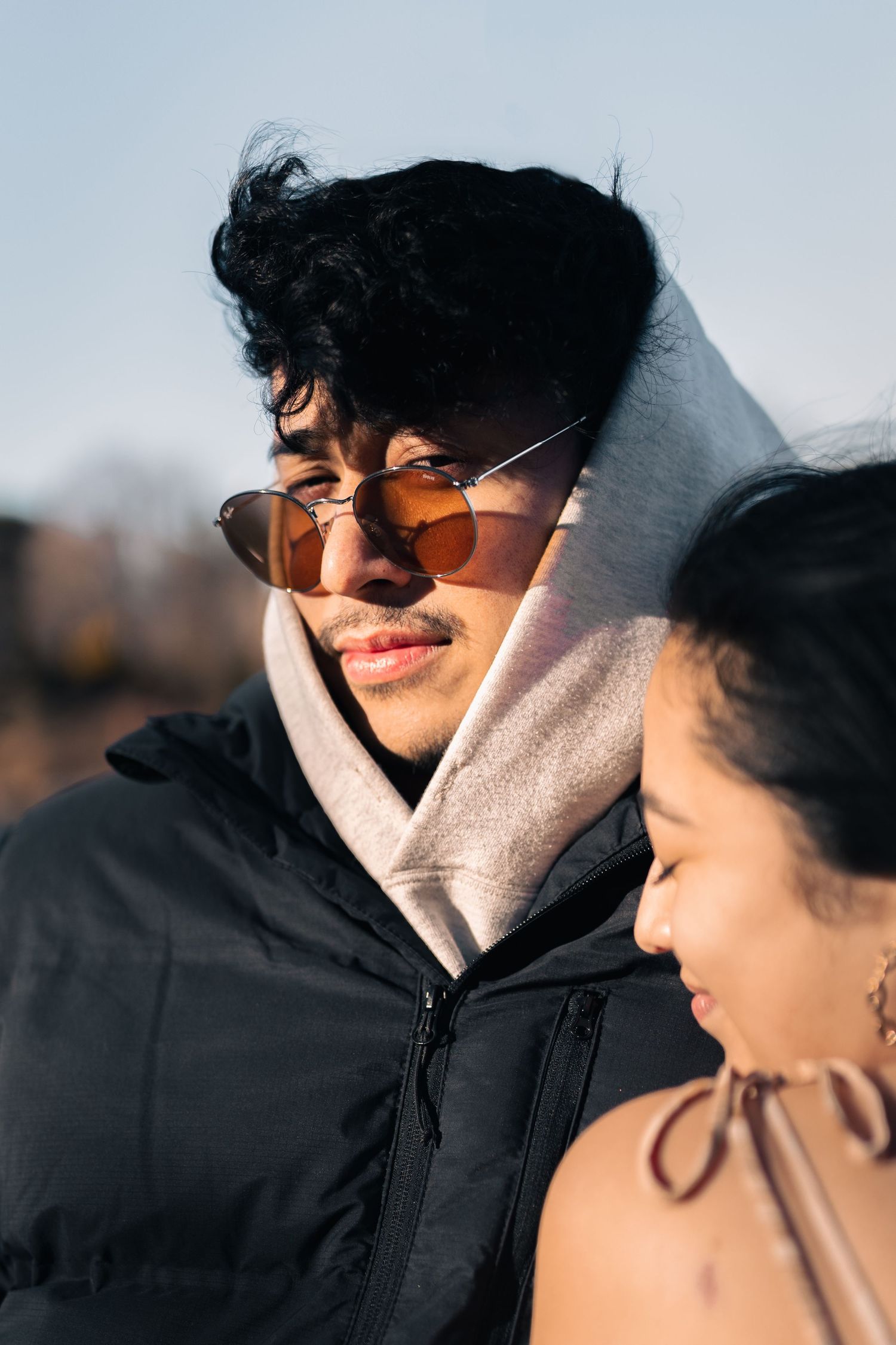Young man wearing orange tinted sunglasses and a hoodie under a black puffer jacket in bright sunlight.