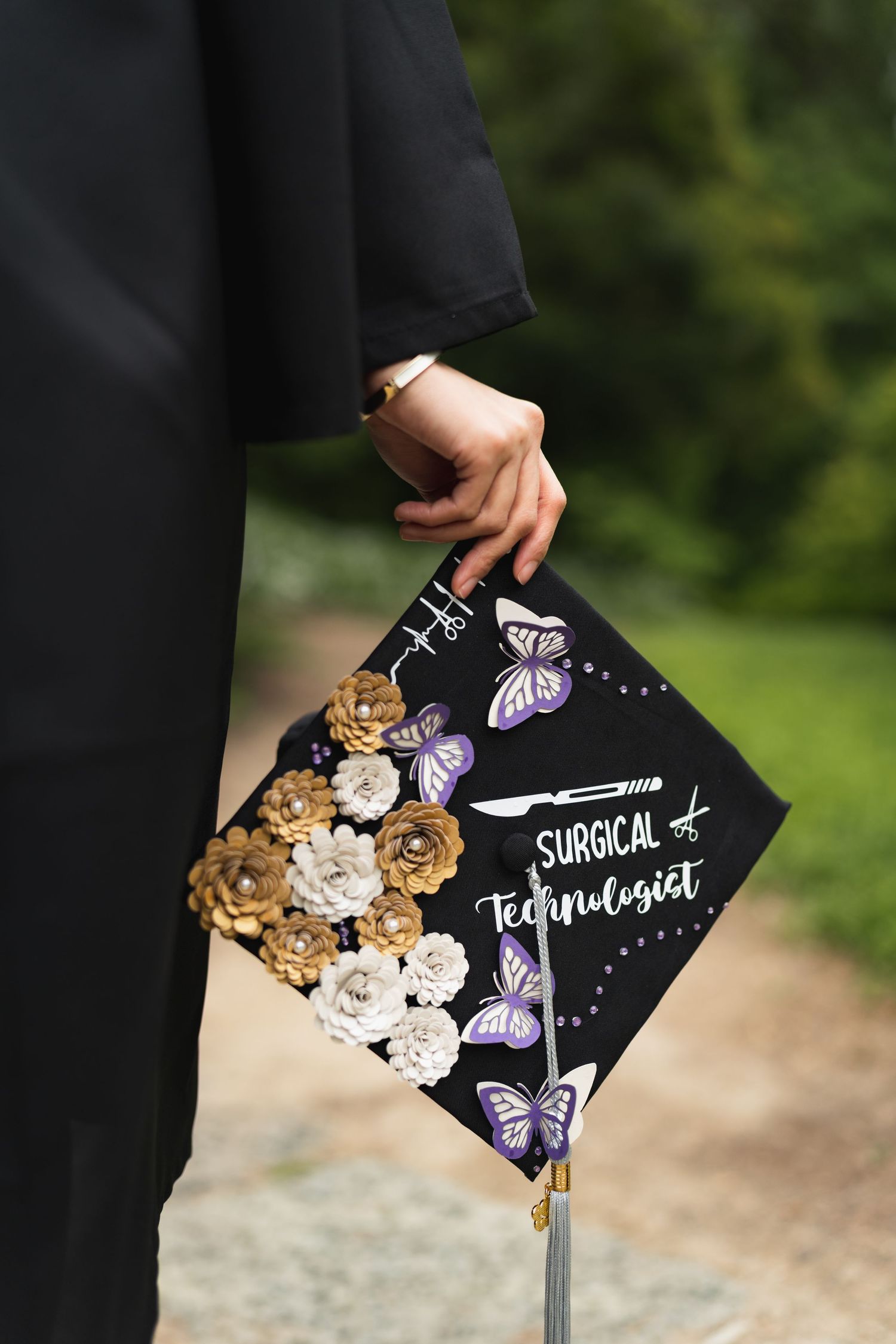 Decorated graduation cap reading Surgical Technologist adorned with purple butterflies and gold paper flowers.