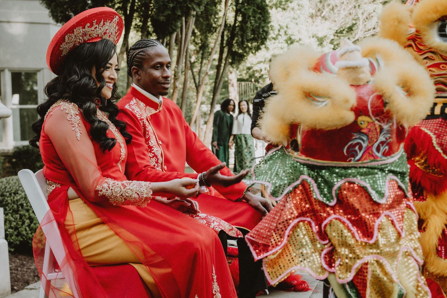 Couple in traditional Vietnamese red ao dai attire seated during a lion dance ceremony at their multicultural wedding.