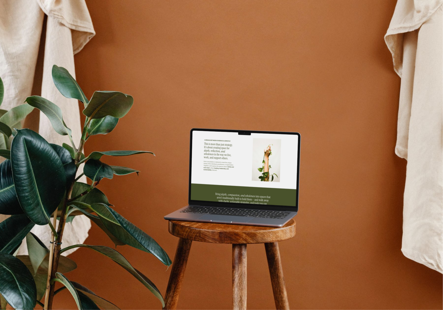 A laptop on a wooden stool displays a green website against a warm terracotta wall with a plant nearby.