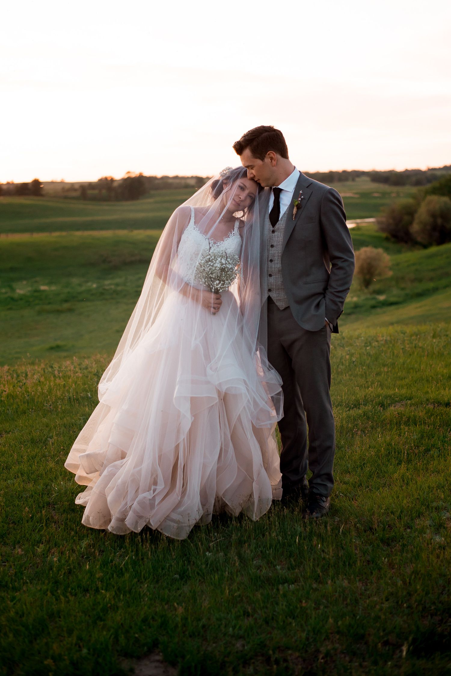 A romantic wedding couple embraces on a grassy hilltop during golden hour, with the bride wearing a flowing blush gown.