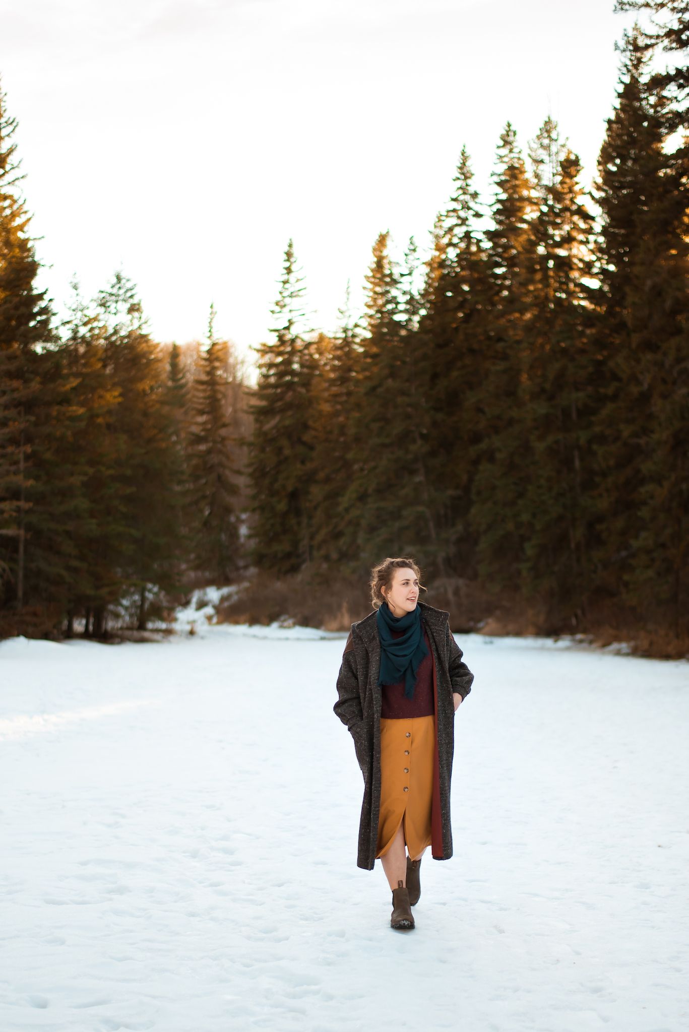 Person in a long dark coat and orange skirt walks through a snowy forest path at sunset.