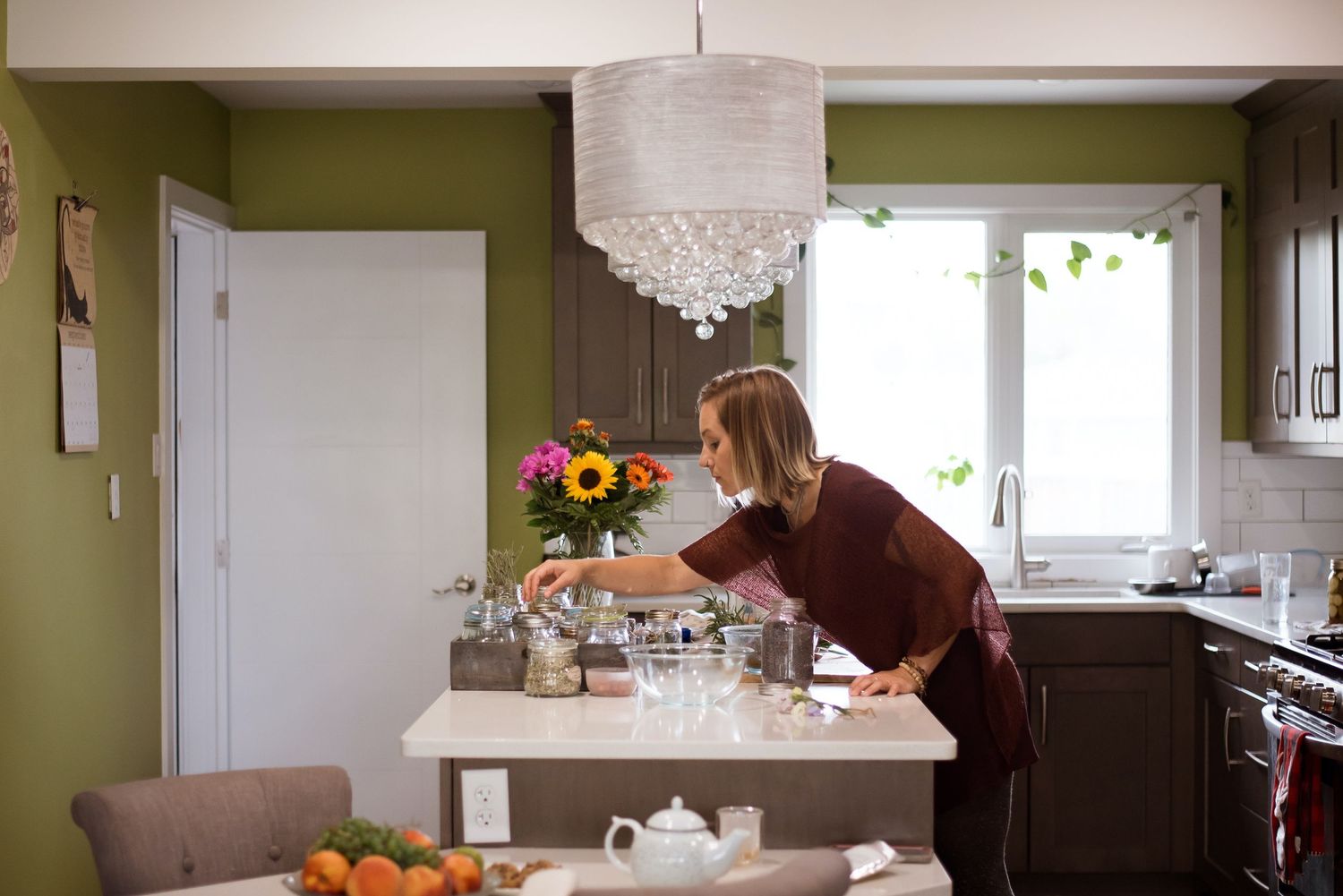 Woman arranging flowers on a kitchen island in a green-walled kitchen with a chandelier overhead.