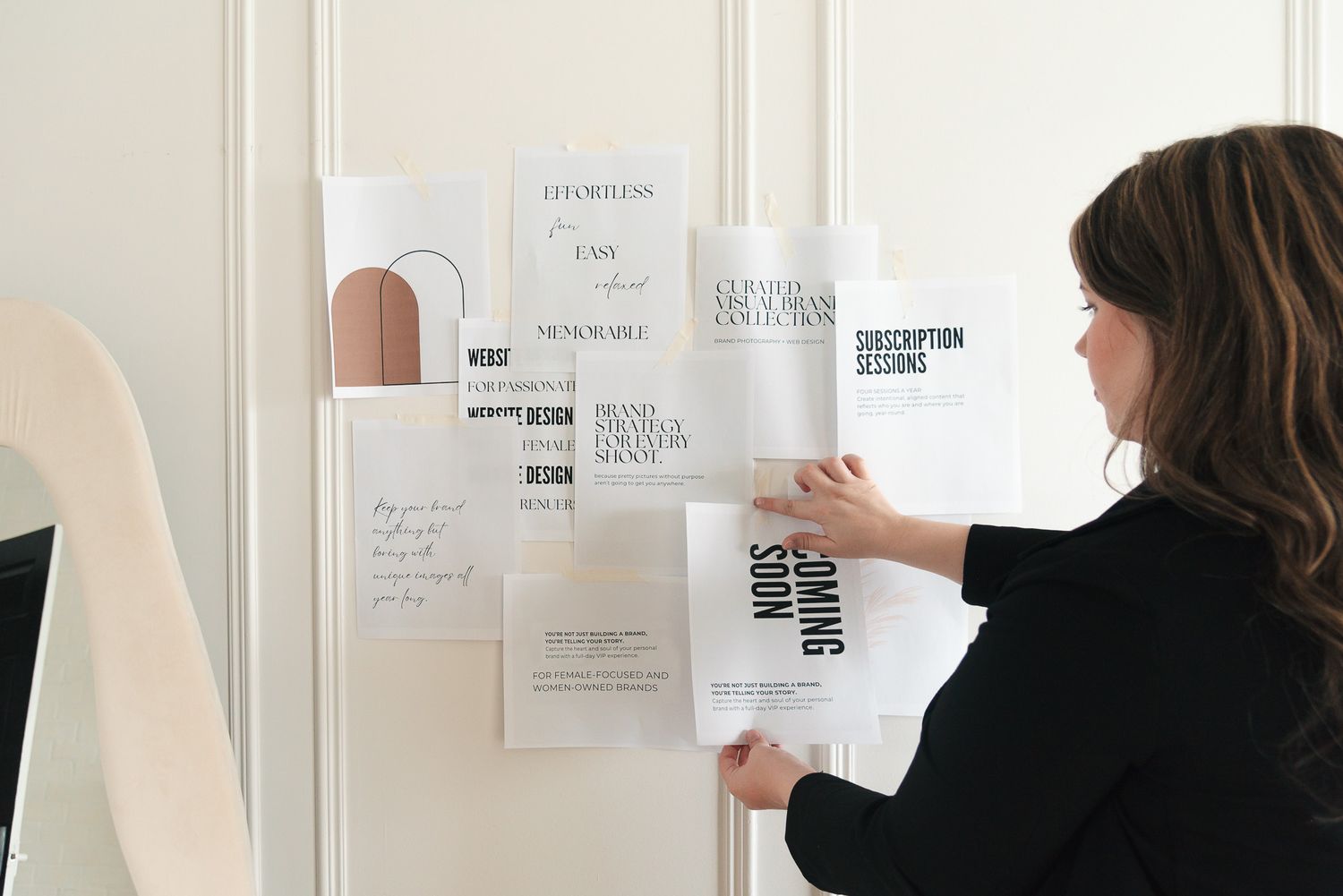 Person in black attaching papers to a white wall displaying design layouts and typographic elements.
