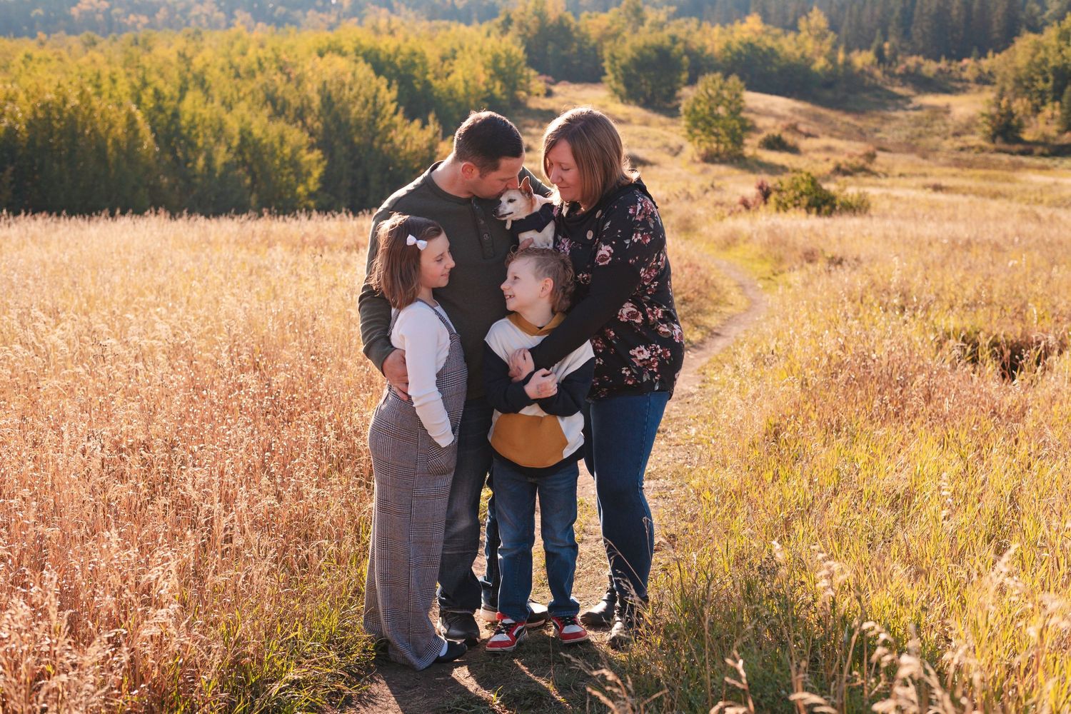 A family embraces together in a sunlit autumn field with golden grass and changing foliage in the background.