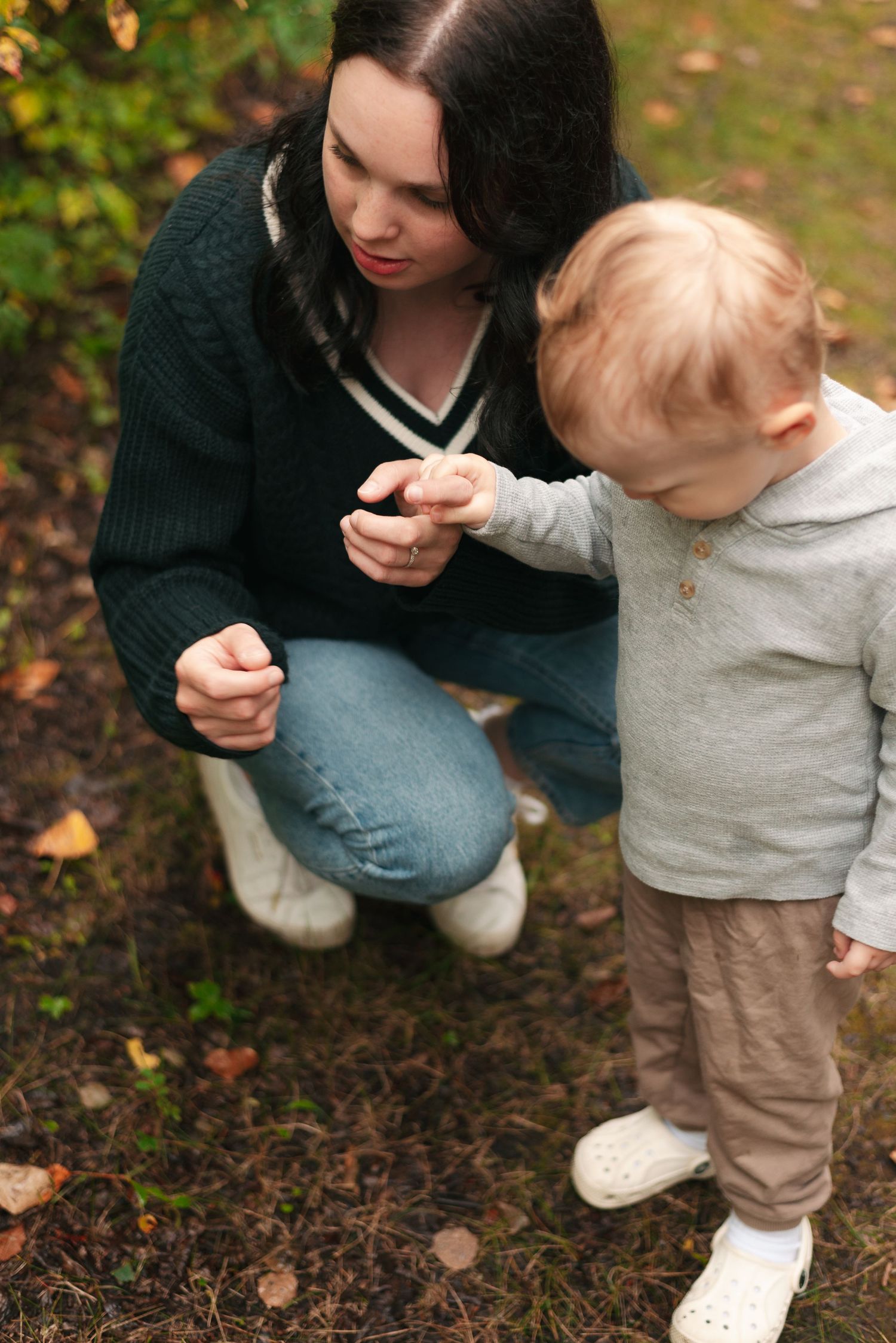 Parent and child kneel together outside examining something in the dirt among scattered autumn leaves.