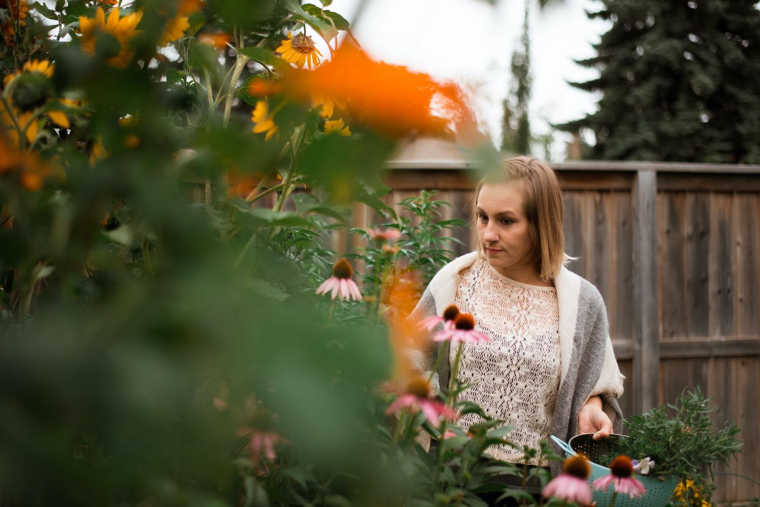 Young blonde woman tending to a colorful garden with pink and orange flowers beside a wooden fence.