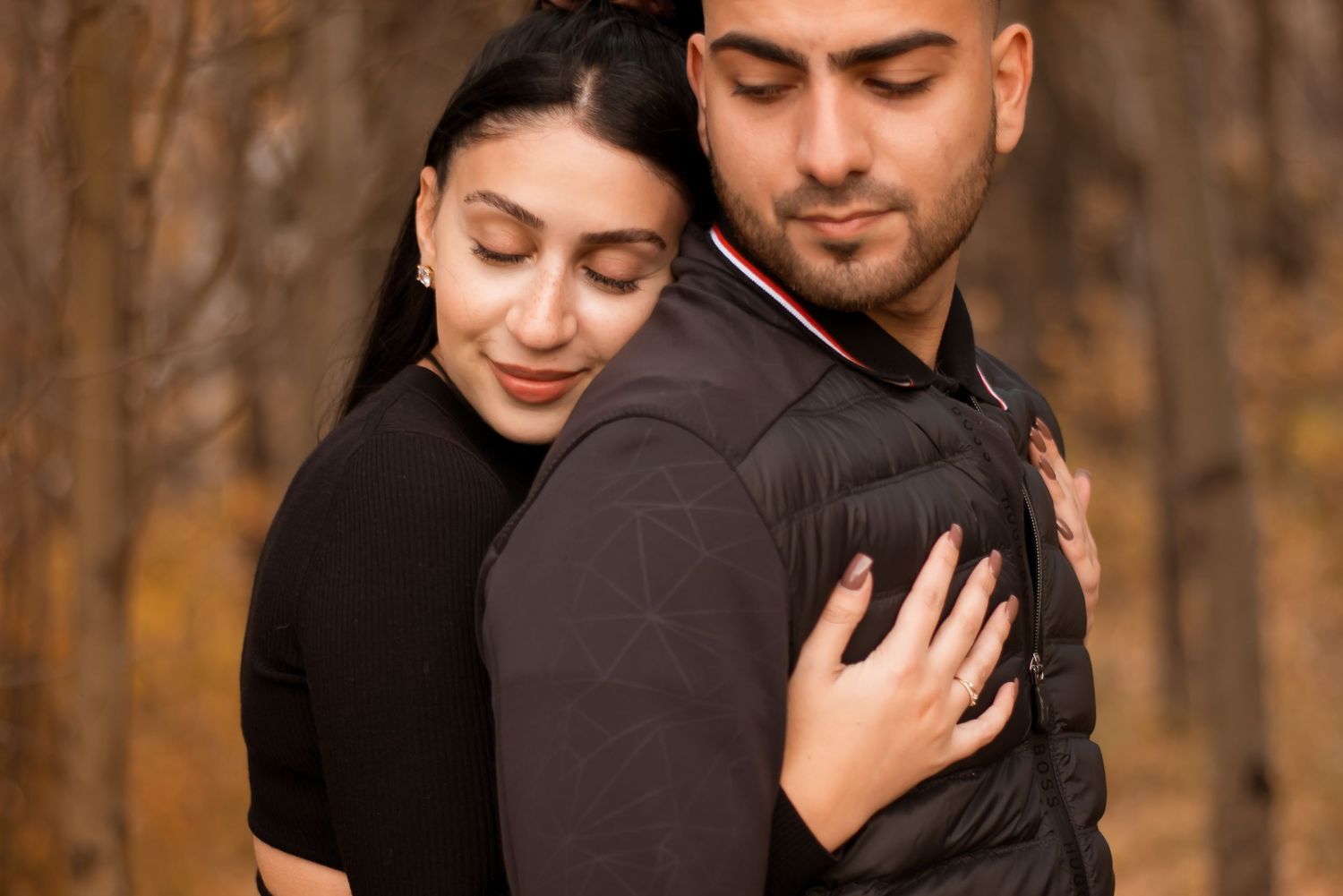 A tender embrace between a couple wearing dark sweaters against an autumn forest backdrop.