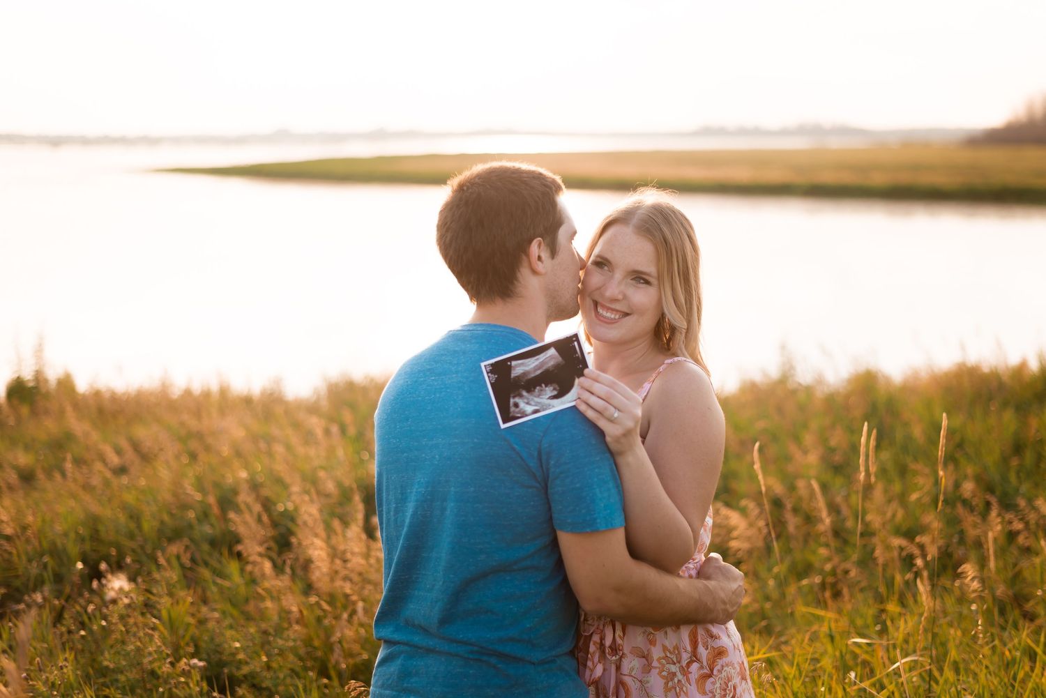 Couple embraces while holding a phone during golden hour by a waterfront with tall grass in the foreground.