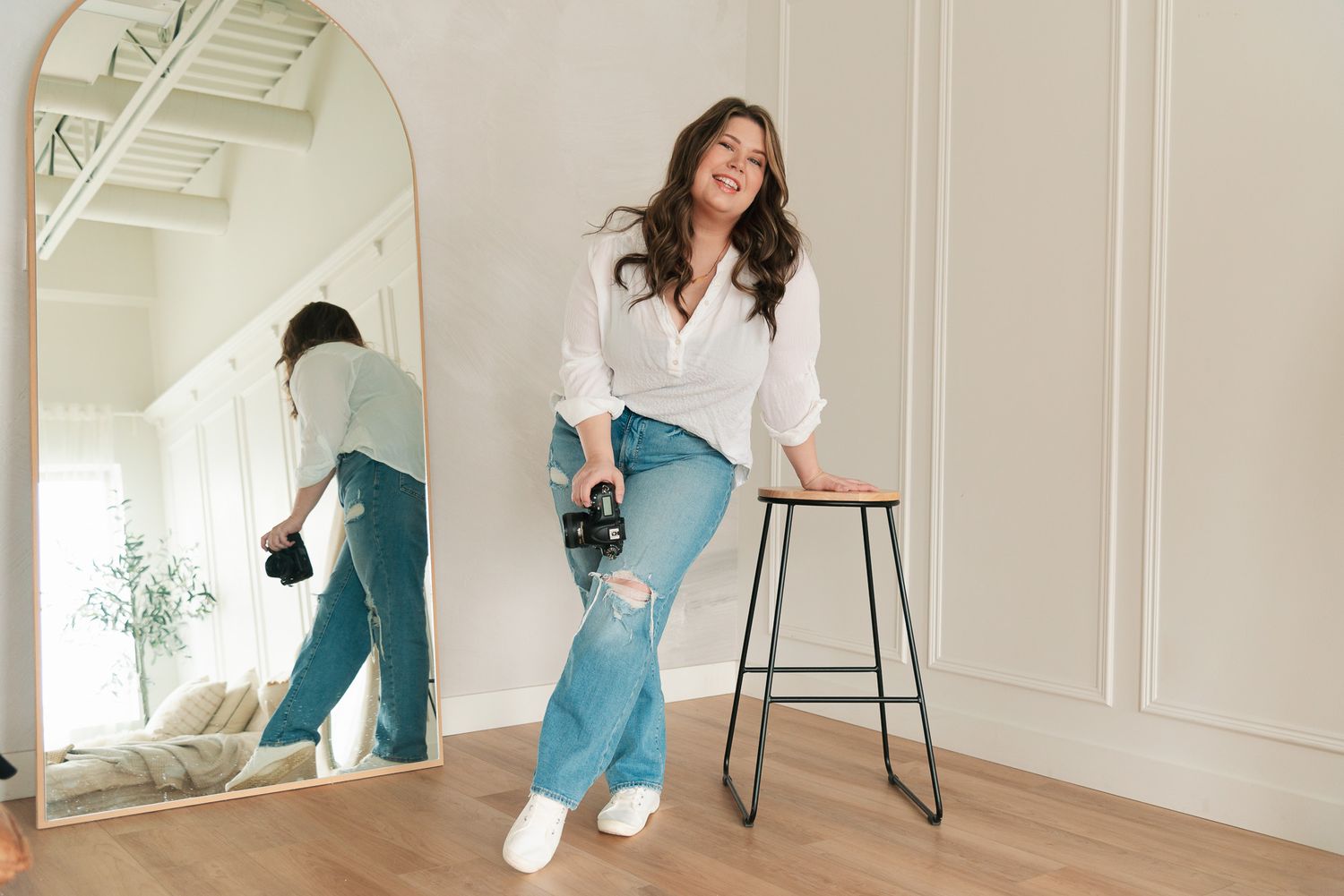 A person wearing a white button-down shirt and light blue jeans poses casually next to a bar stool in a bright room.