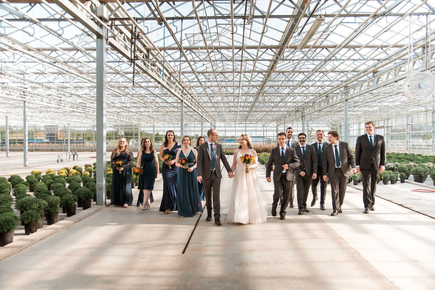 Wedding party walking together in a modern greenhouse structure with glass ceiling and plants lining the sides.
