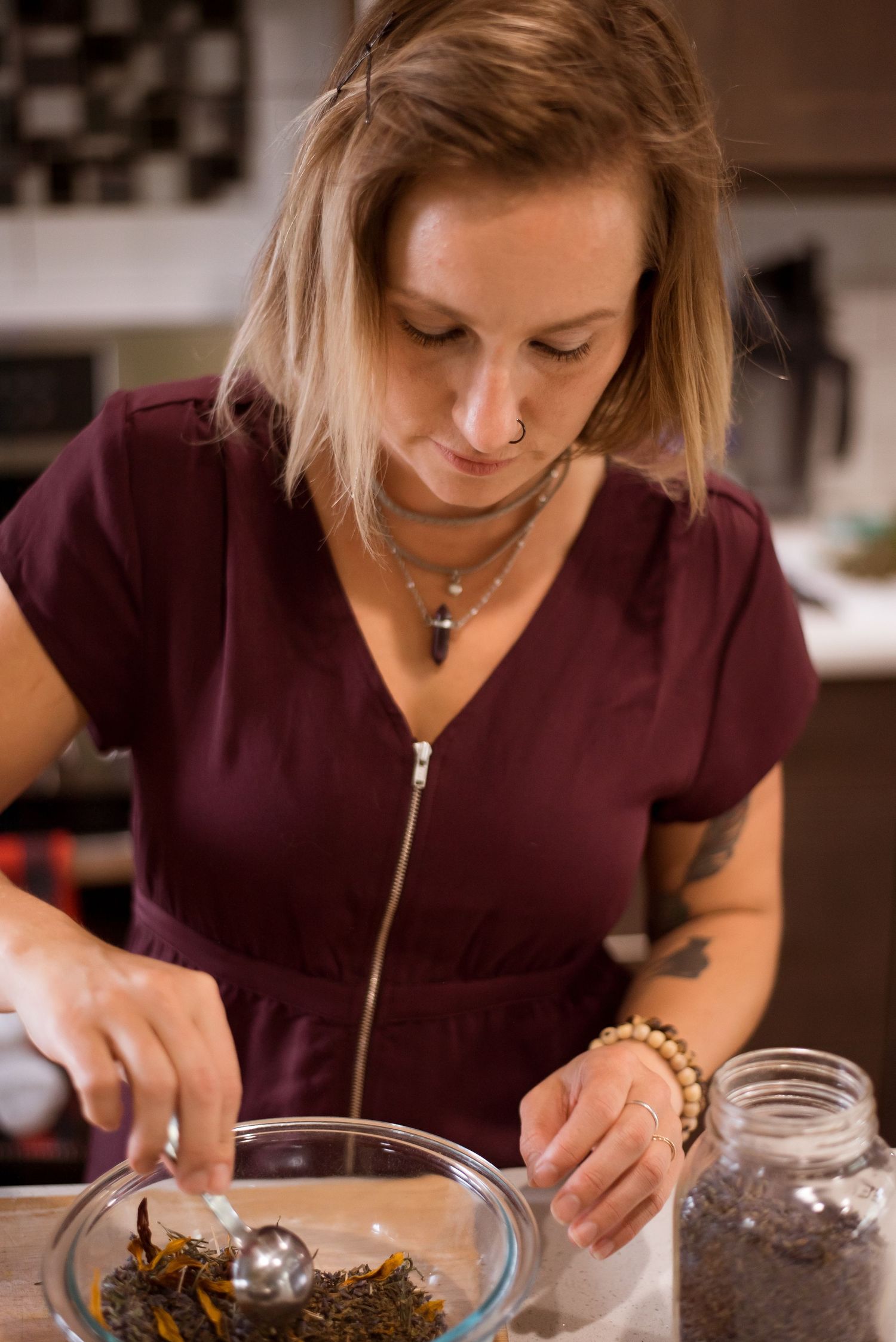 Woman in maroon dress preparing loose leaf tea with a strainer ball in a glass bowl in a kitchen.