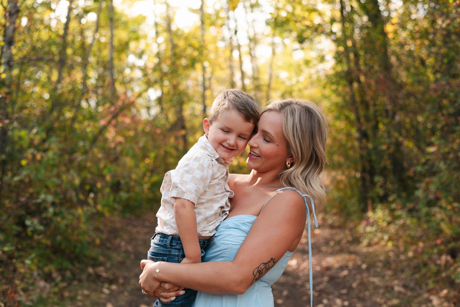 A woman holds a smiling toddler boy in a sunlit autumn forest, both looking joyful in casual outfits.