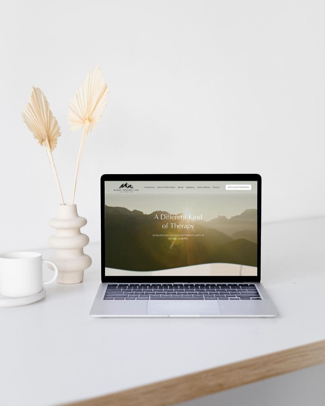 Minimalist desk setup with laptop displaying mountain scenery and white ceramic decor against light background.