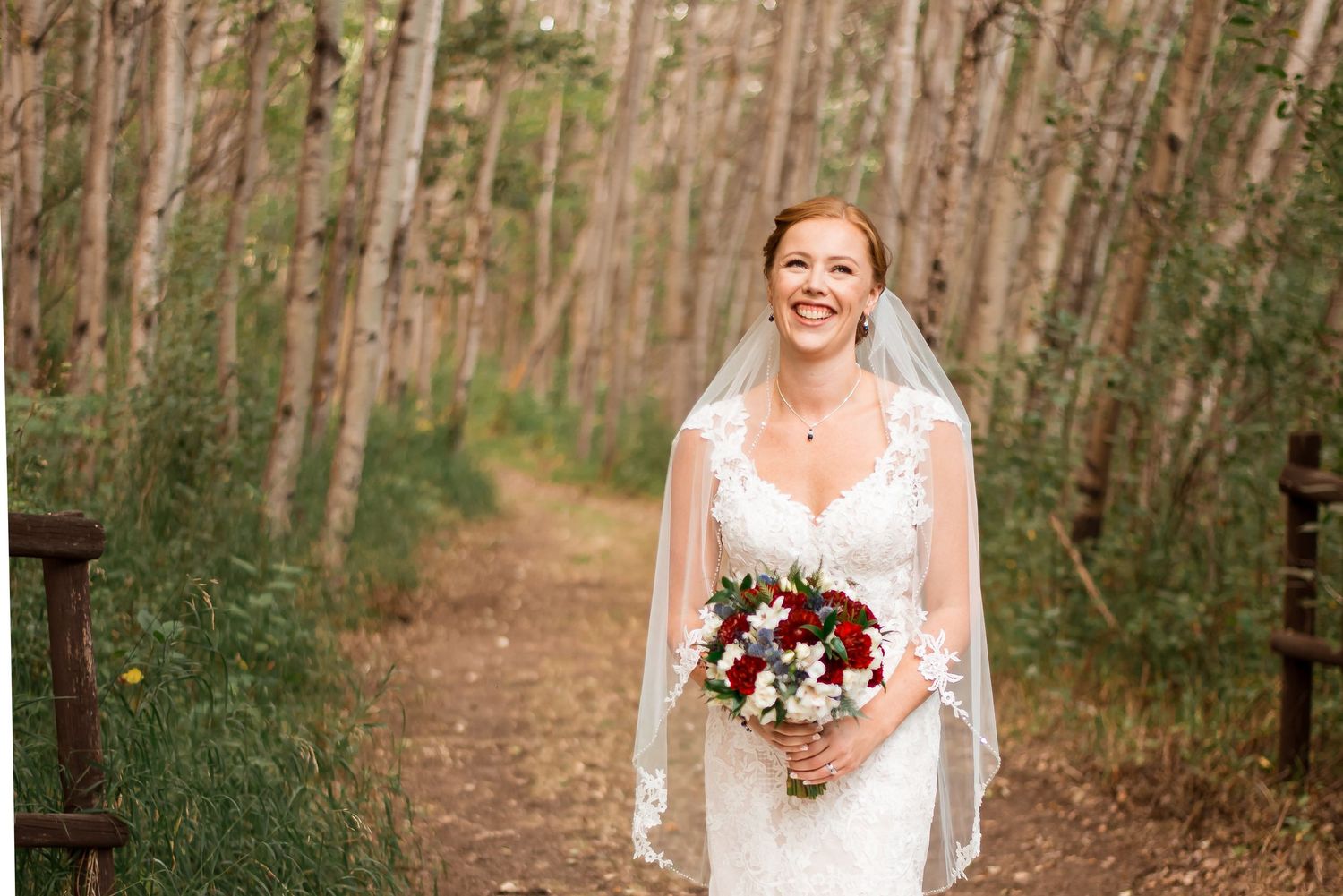 A smiling bride wearing a lace white dress and veil holds a colorful bouquet while standing on a forest trail.