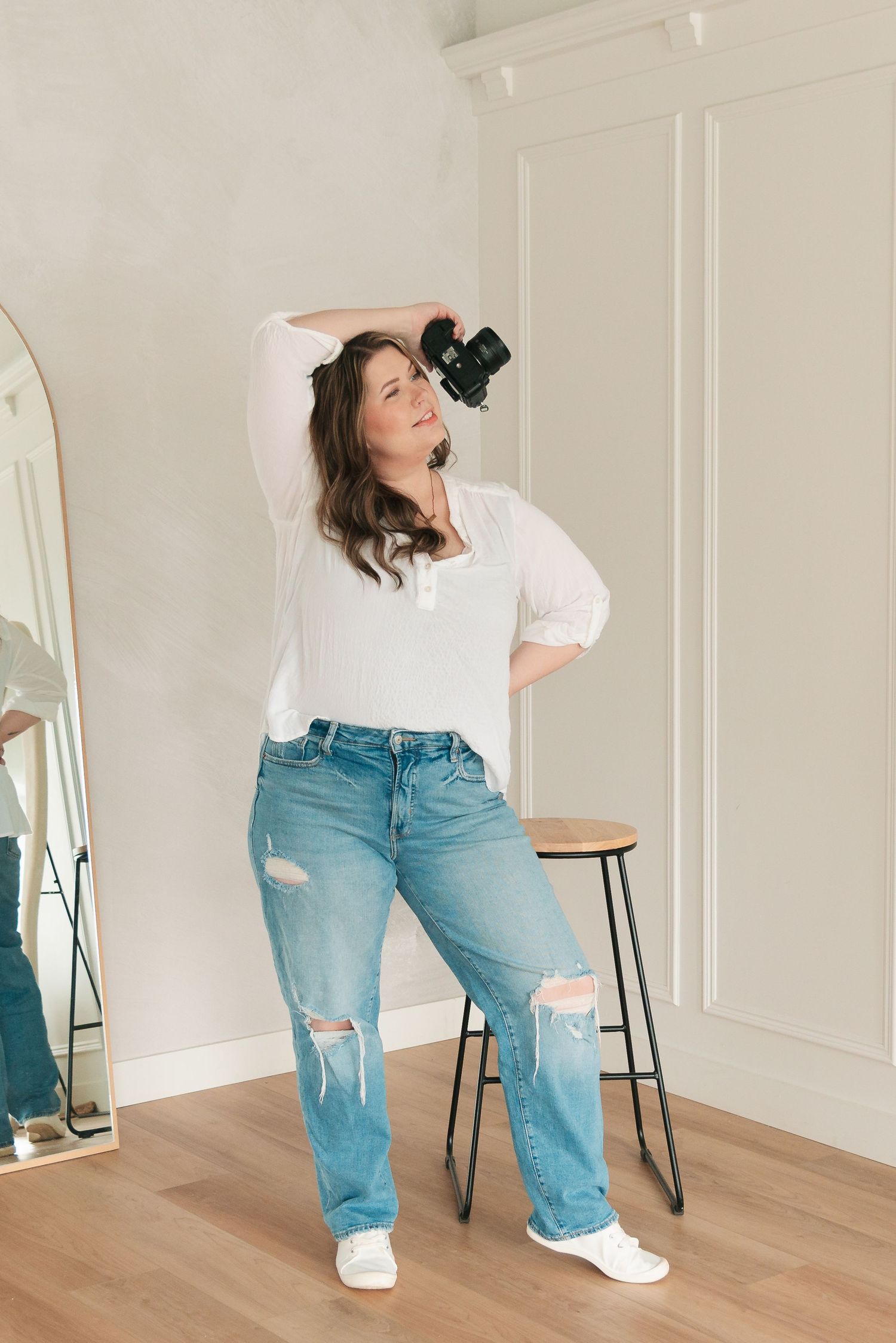 Person in casual attire with white blouse and distressed blue jeans poses playfully in minimalist room with mirror.
