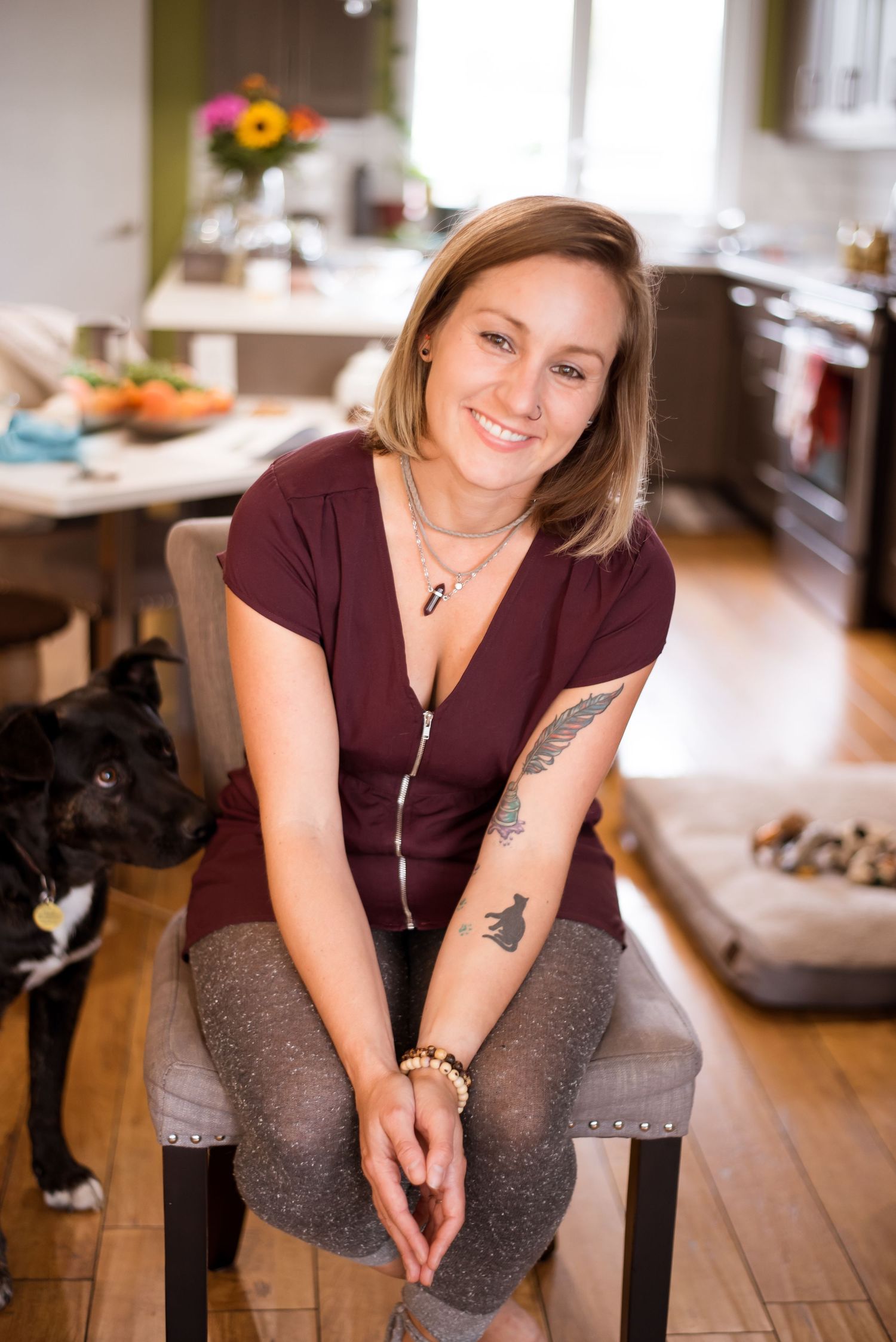 Woman in burgundy shirt sits on chair in cozy kitchen with dog beside her.