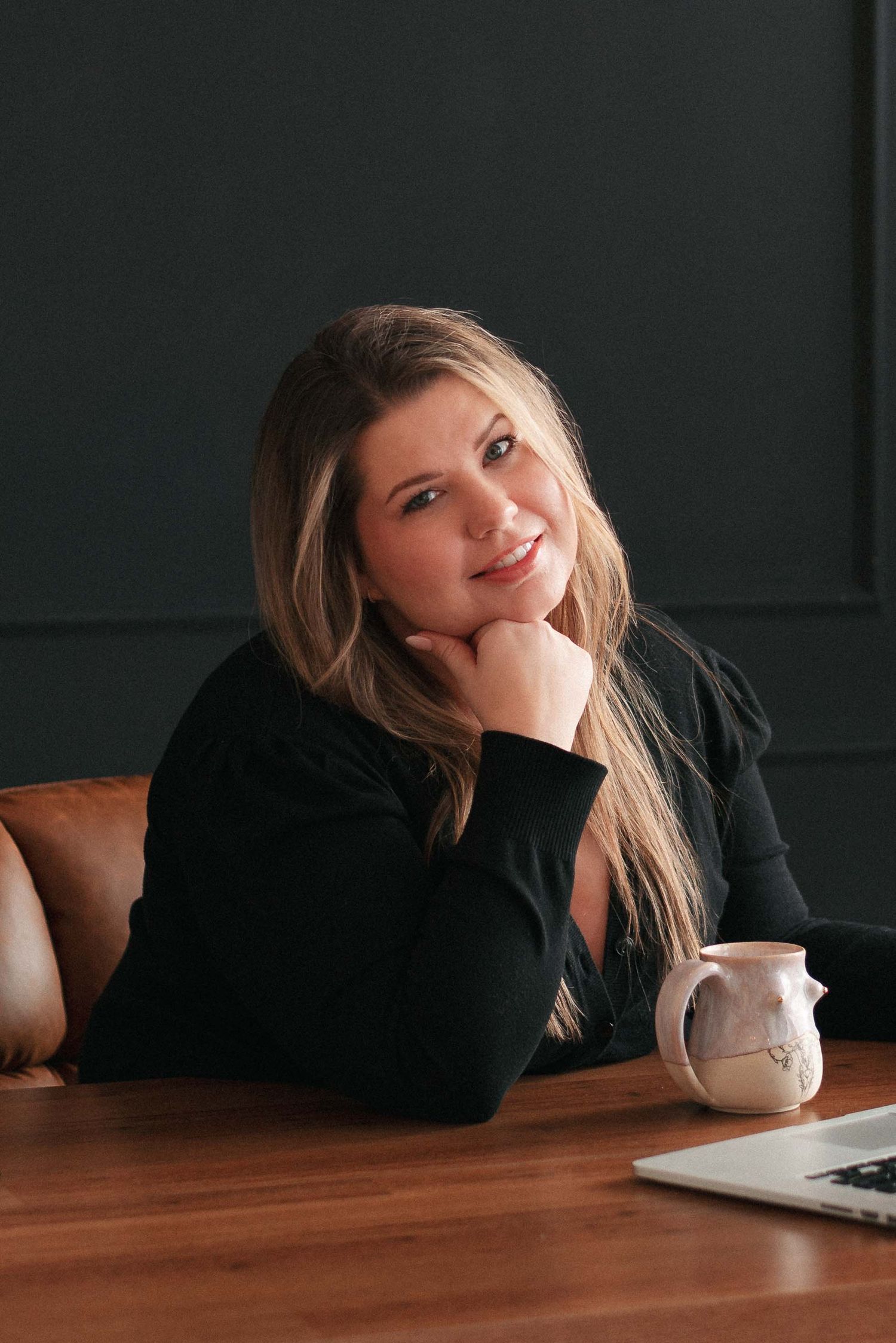Person in black sweater rests head on wooden table with coffee cup in moody indoor setting.