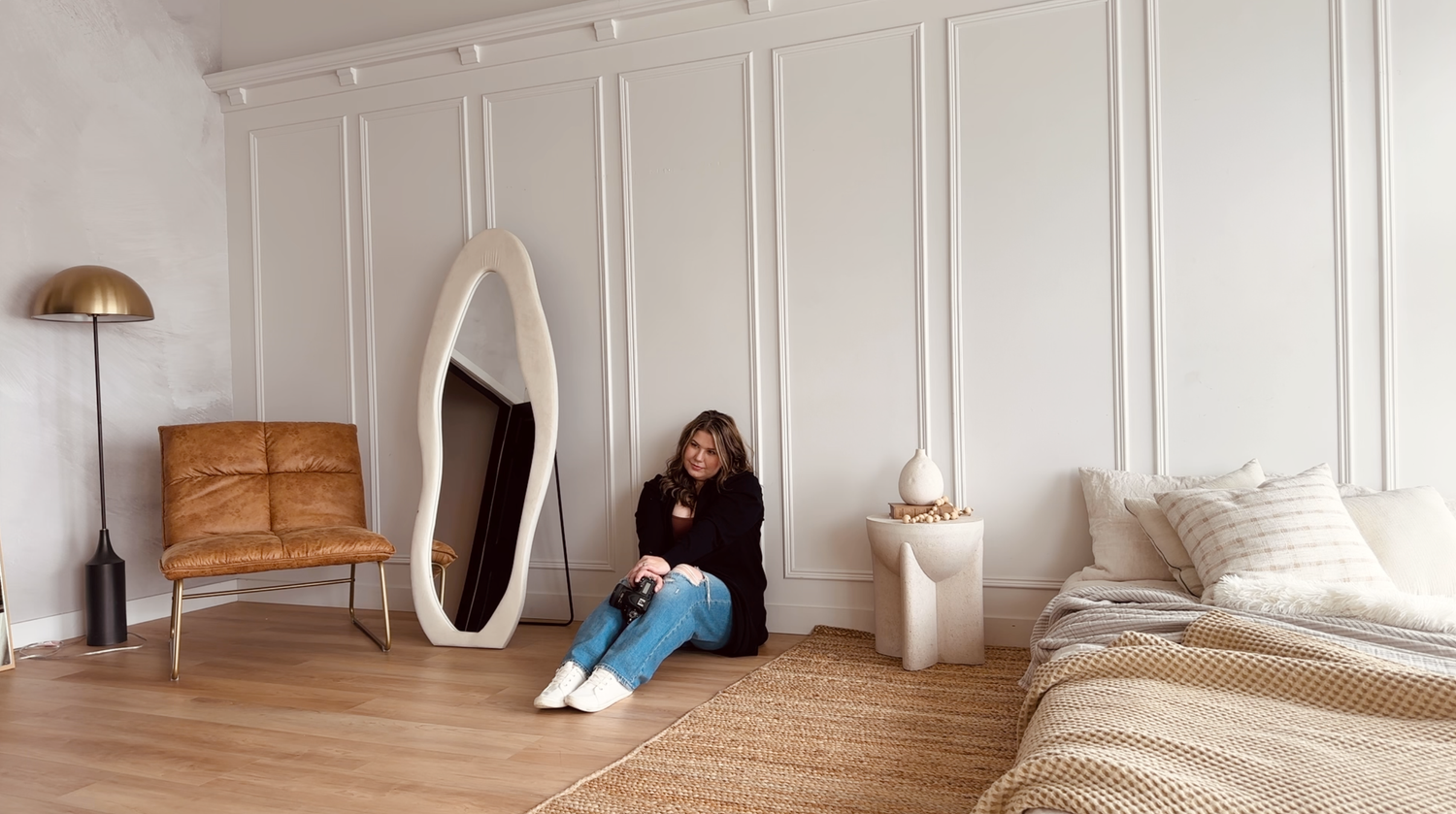 A room with wooden floors featuring a freestanding oval mirror, tan chair, bed, and minimalist decor against white walls.