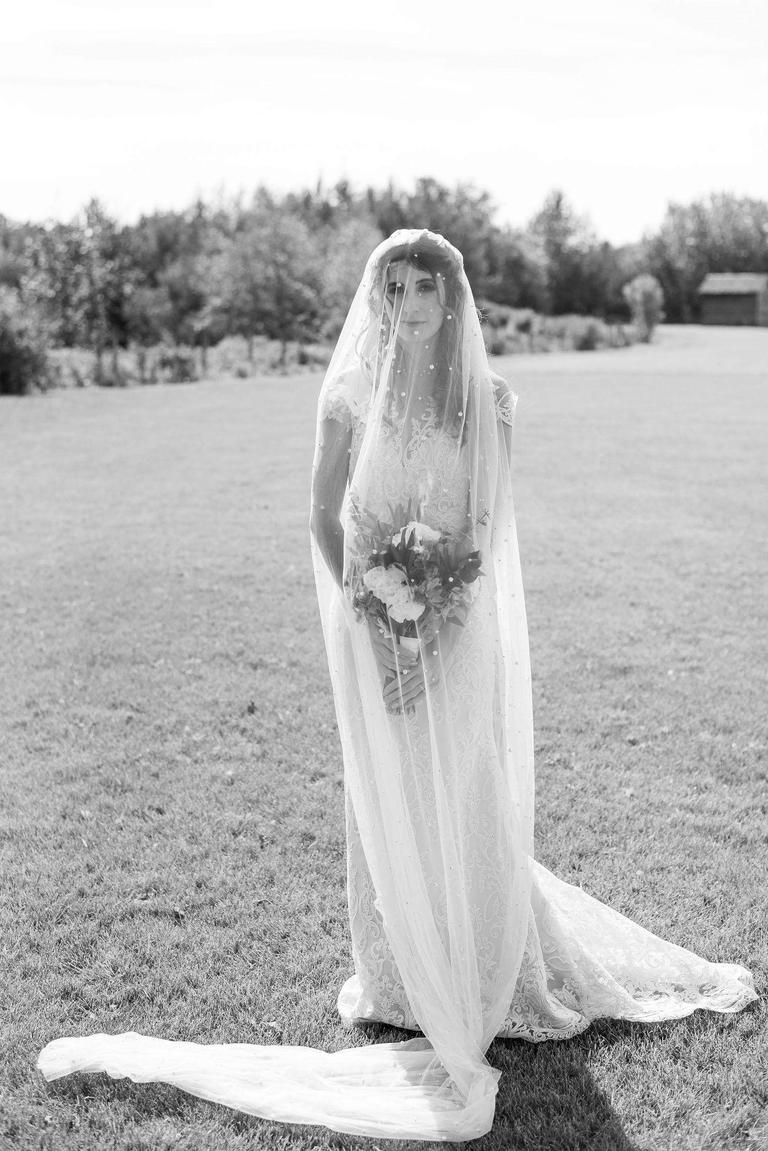 Black and white photo of bride in flowing lace wedding gown and long veil holding bouquet while standing on grass.